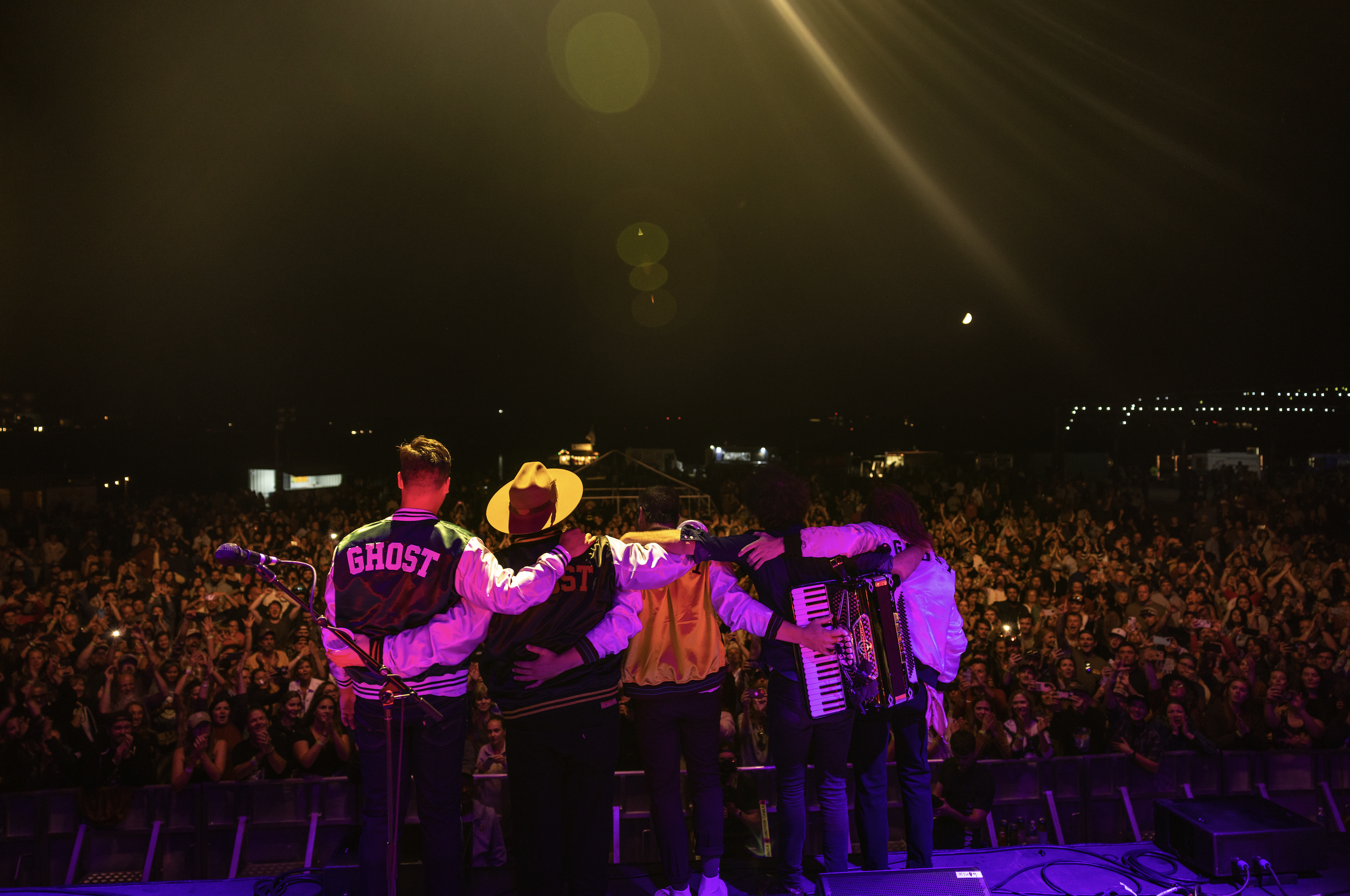 Musicians stand on stage with their backs to the camera, facing a large audience at a concert or festival during nighttime. The crowd is enjoying the performance, and city lights are visible in the background, with a dark night sky above.