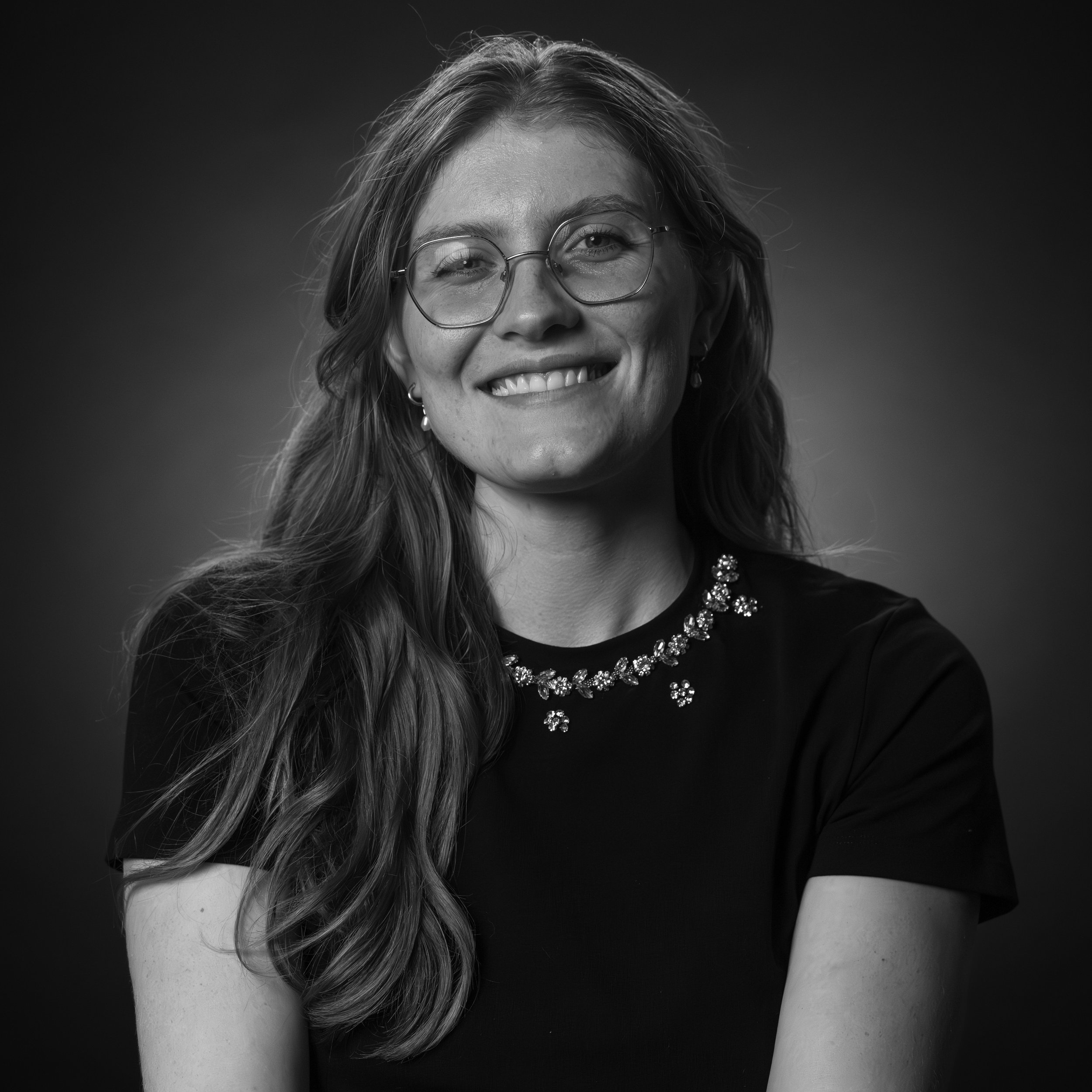 Black and white portrait of a smiling woman with long wavy hair, glasses, and a necklace, wearing a short-sleeved top.