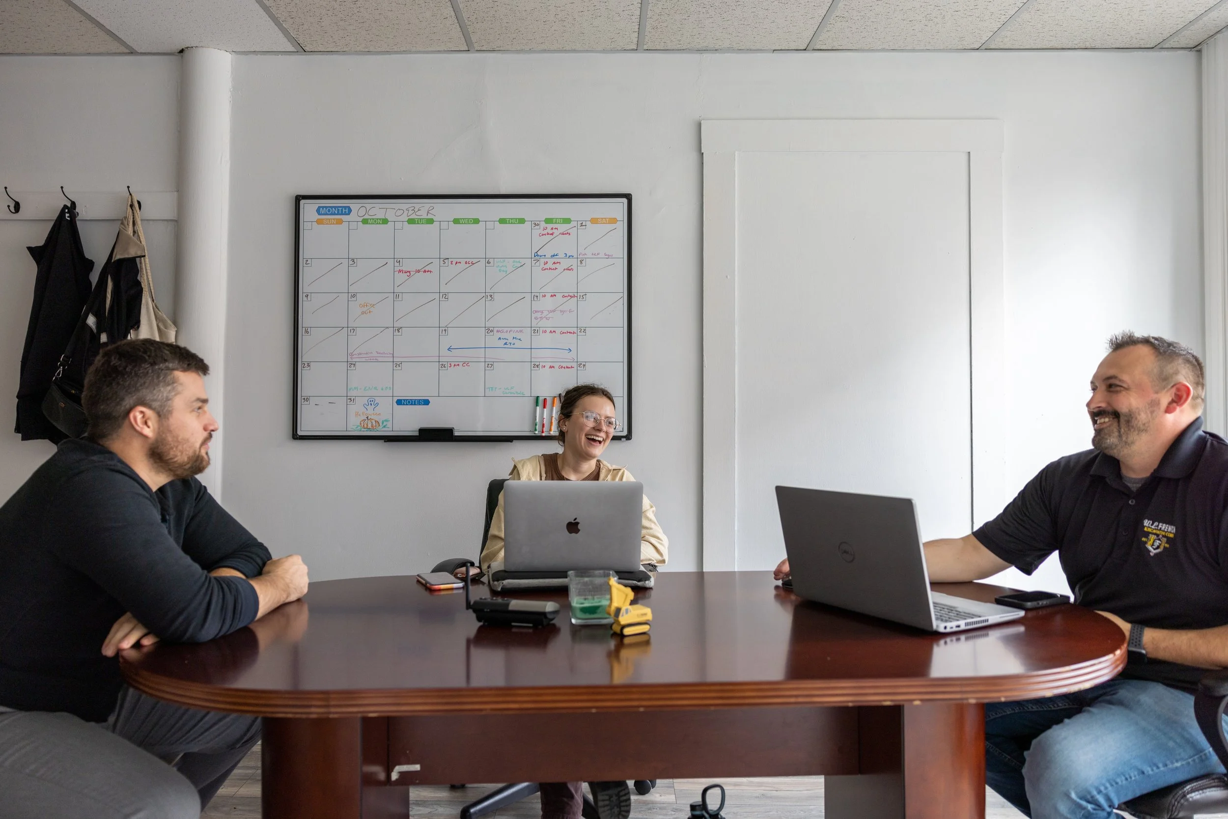 Three people sitting at a conference table in a meeting room, with laptops, notebooks, and a whiteboard in the background. They are engaged in a discussion, with one woman smiling.