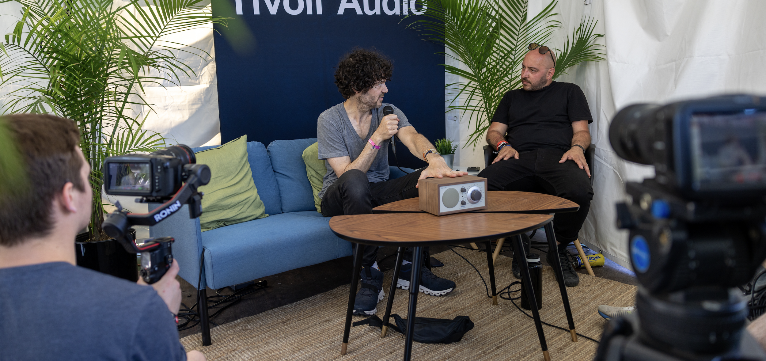 Two men are sitting on a blue couch in a tent, one is holding a microphone and speaking, the other listens. There are two cameras recording the interview, and a person in the foreground is operating one of the cameras. The scene has plants and a blue and white backdrop with part of text visible.