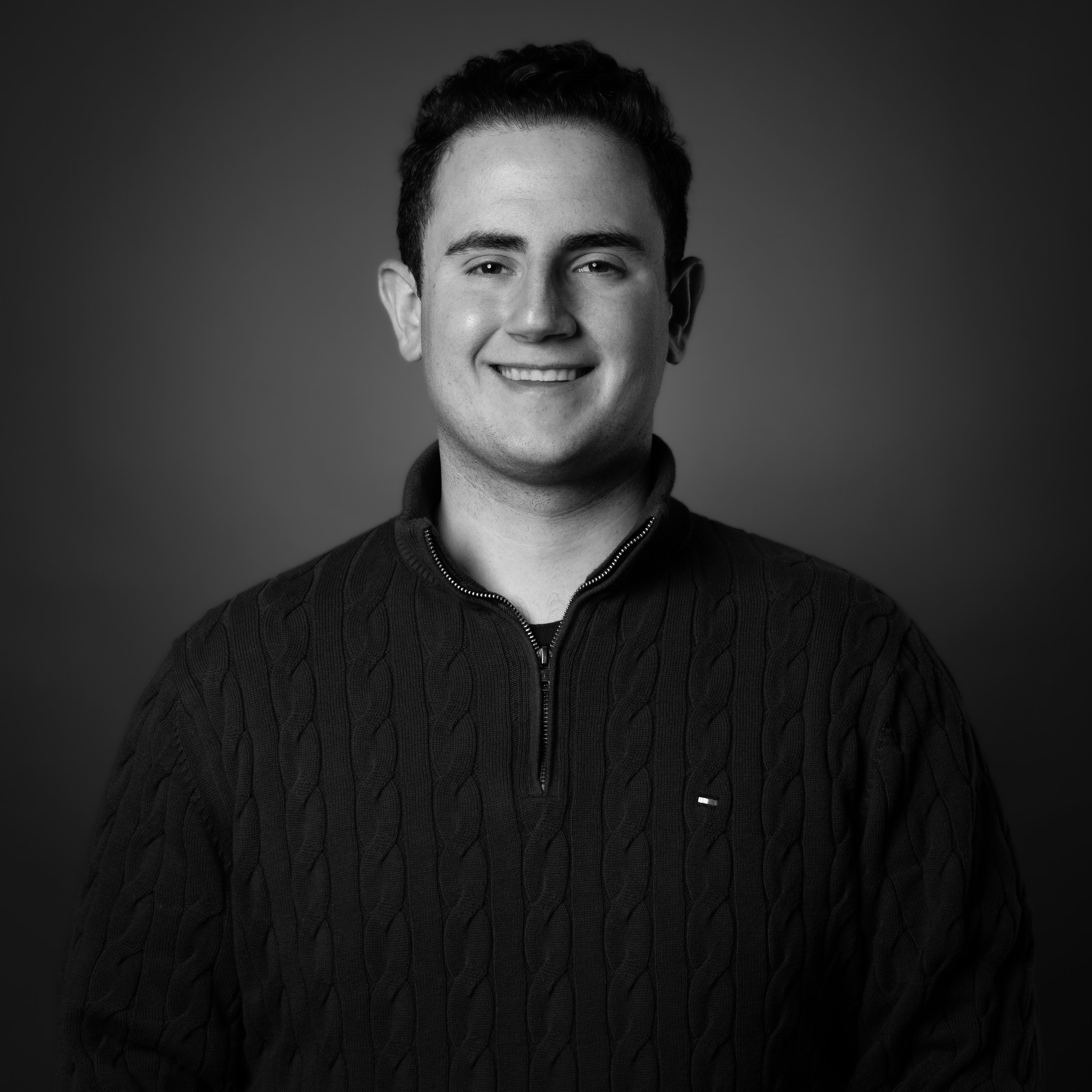 Black and white portrait of a young man smiling, wearing a dark zippered sweater with a textured knit pattern, against a plain background.
