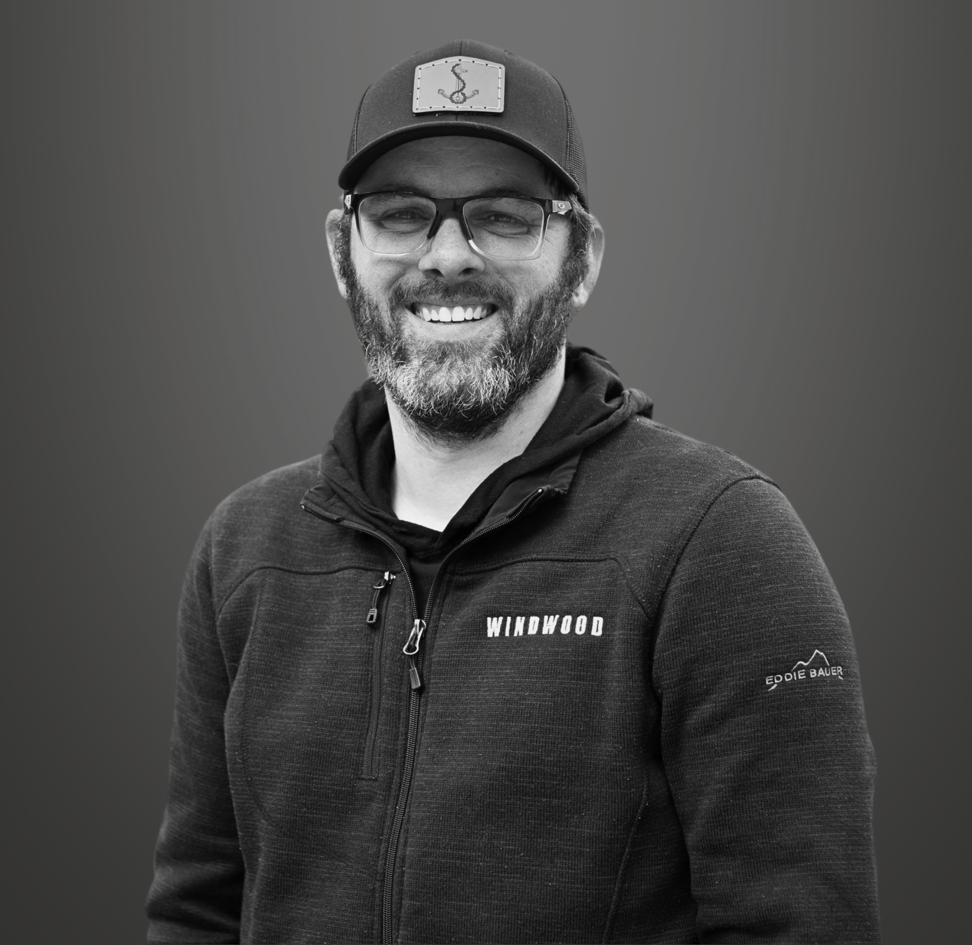 Black and white photograph of a smiling man with glasses, a beard, wearing a baseball cap, a zip-up hoodie with the word 'Windwood' on it, and a logo on the sleeve, standing against a plain background.