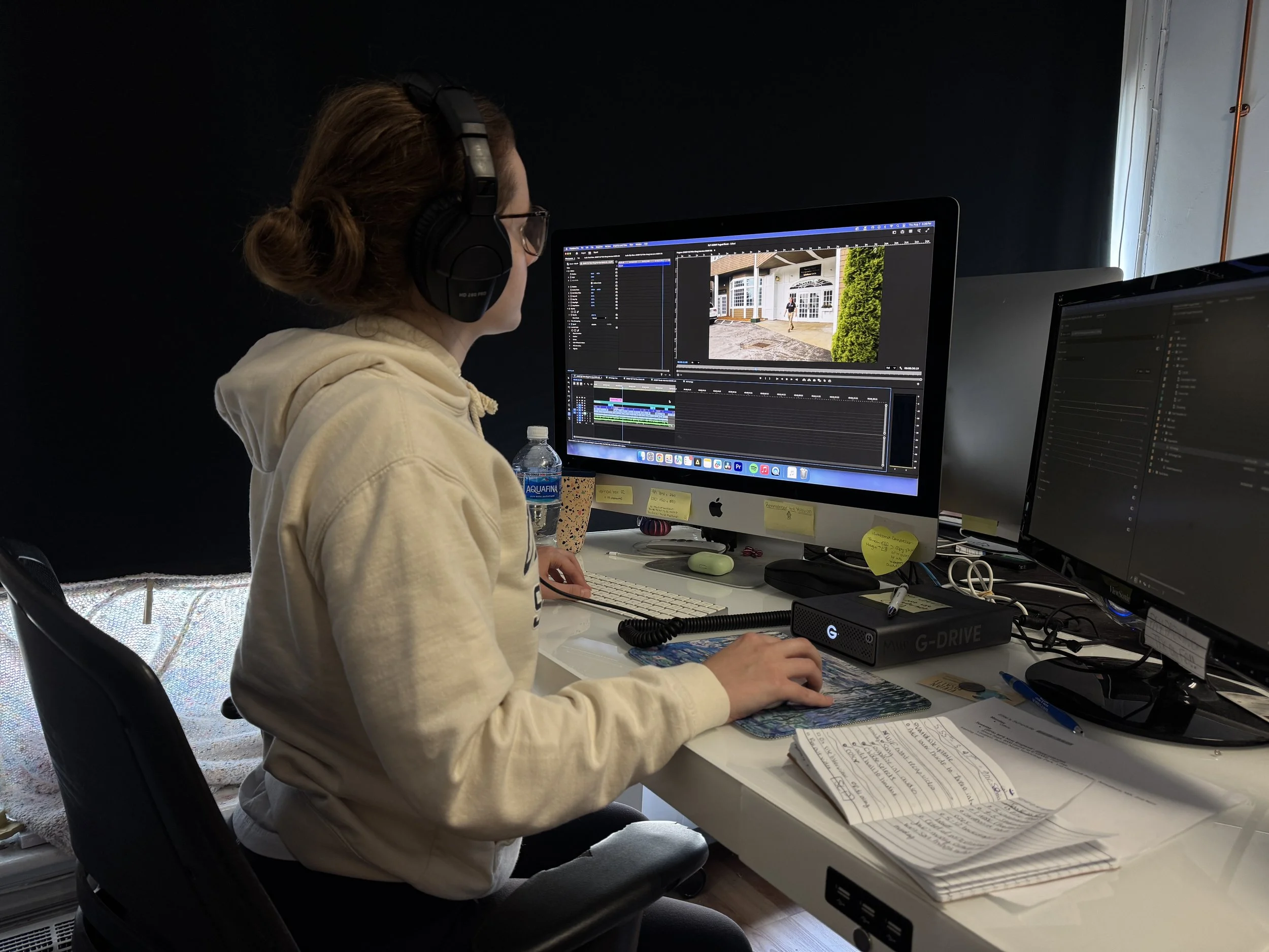 A person with headphones working on video editing at a desk with dual monitors, papers, and a water bottle.