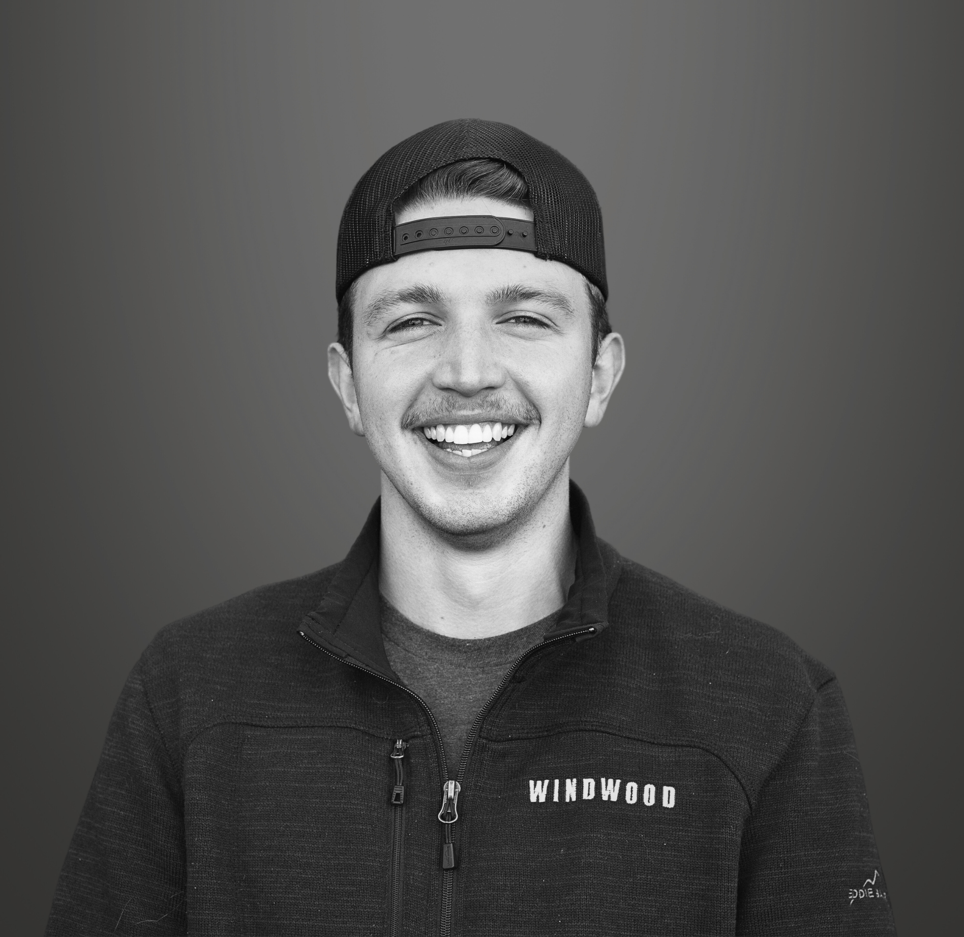 Black and white portrait of a smiling man wearing a backward baseball cap and a dark zip-up jacket with 'Windwood' embroidered on it, against a neutral background.