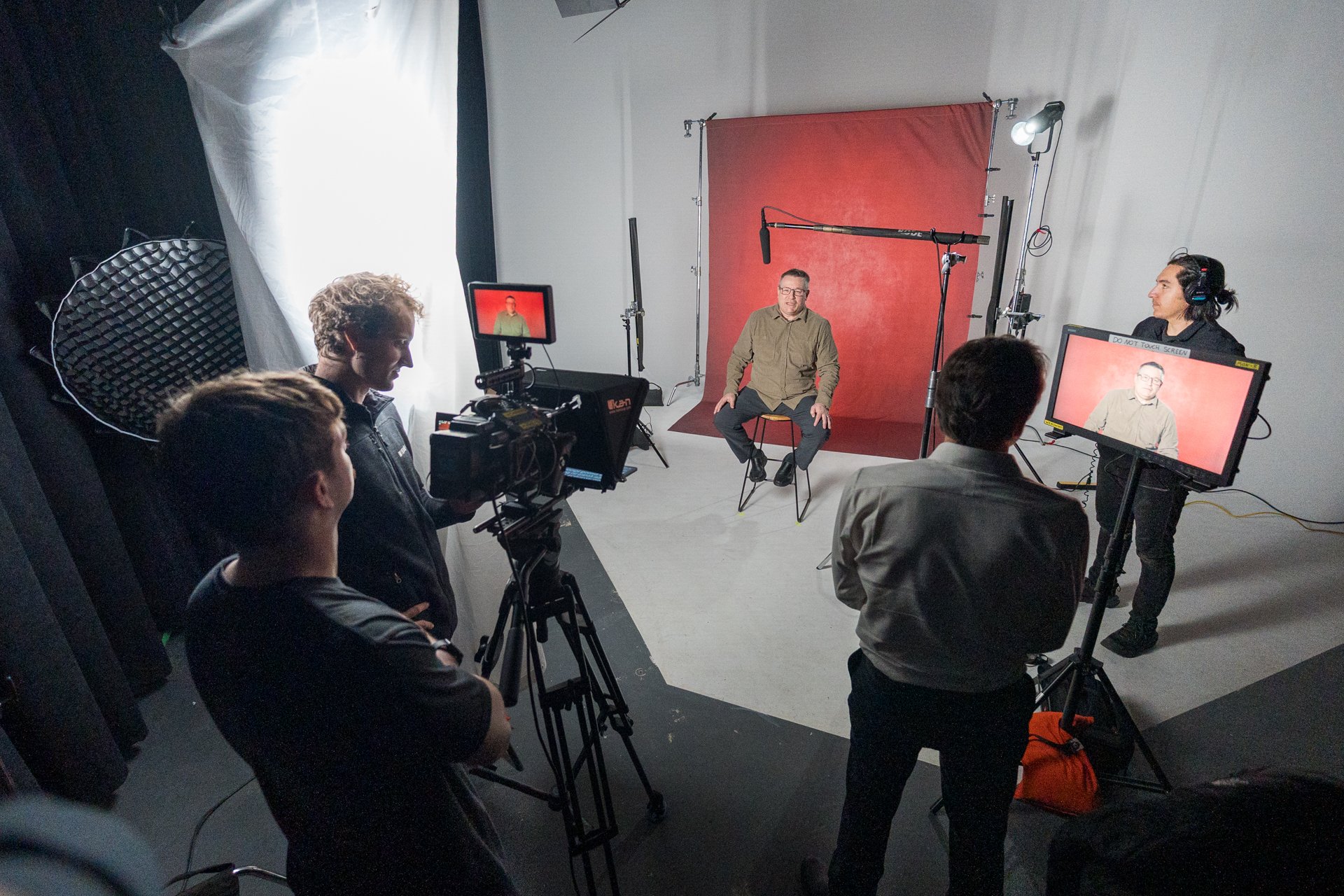 A filming crew is inside a studio setting, capturing a man sitting on a stool in front of a red backdrop. Crew members operate cameras, a boom microphone, and lighting equipment, with a monitor showing the man's live image.