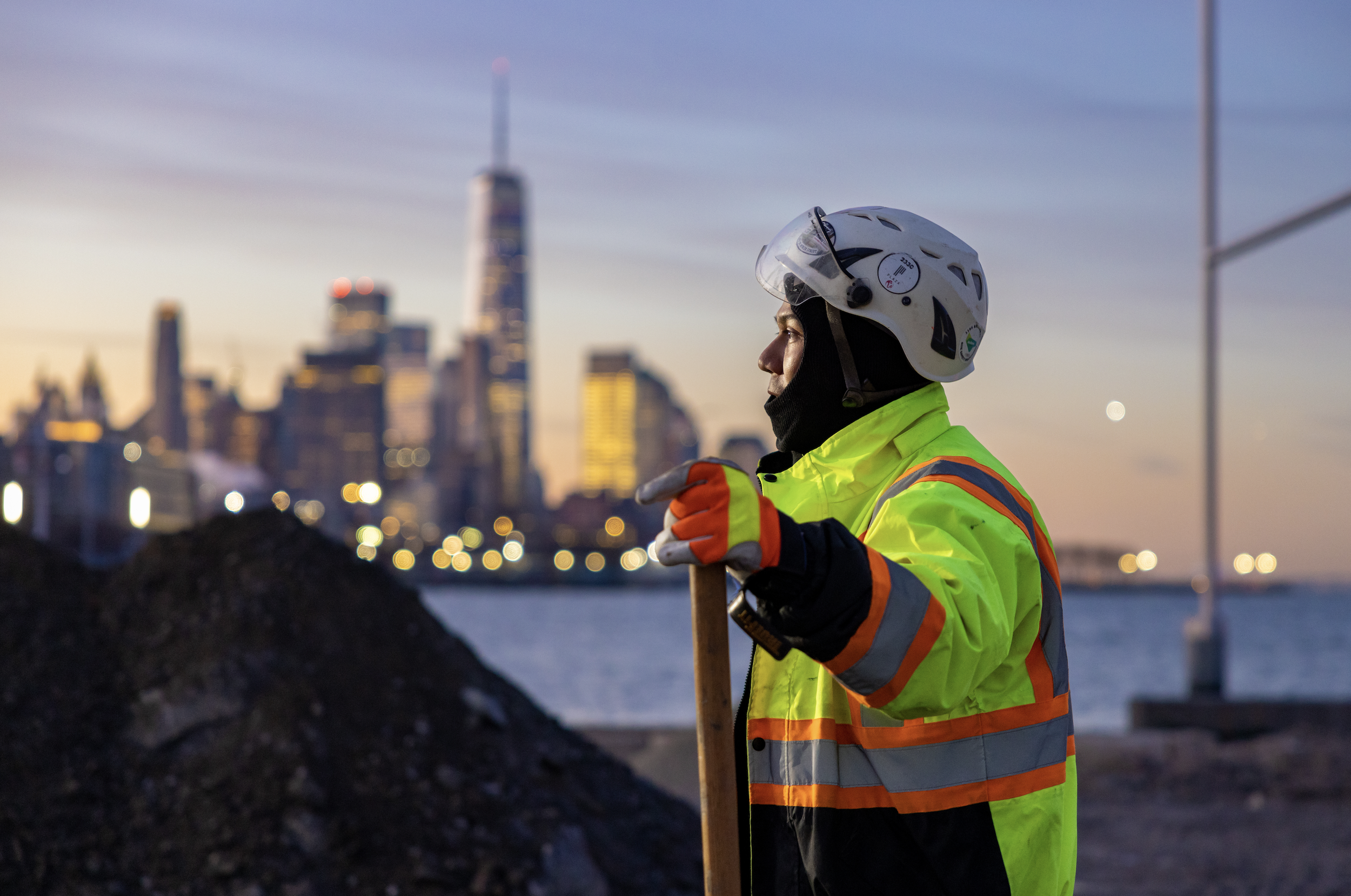 BTS working with Windwood: Construction worker in high-visibility jacket and helmet standing near water, holding a tool with blurred city skyline in the background at sunset.