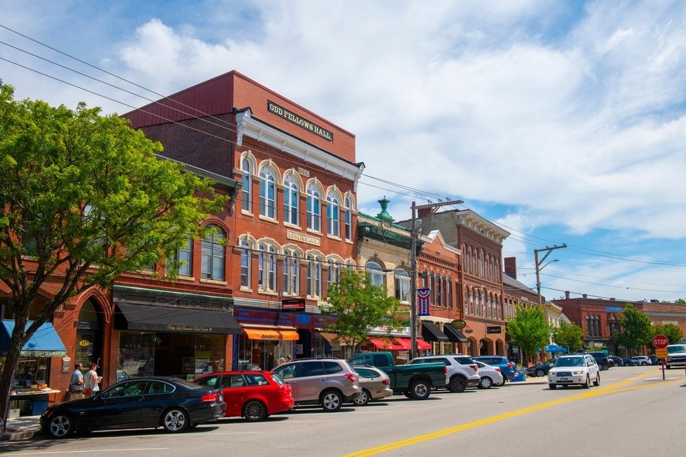 Main street in Concord nh- with historic brick buildings, trees, parked cars, and a few pedestrians under a partly cloudy sky.