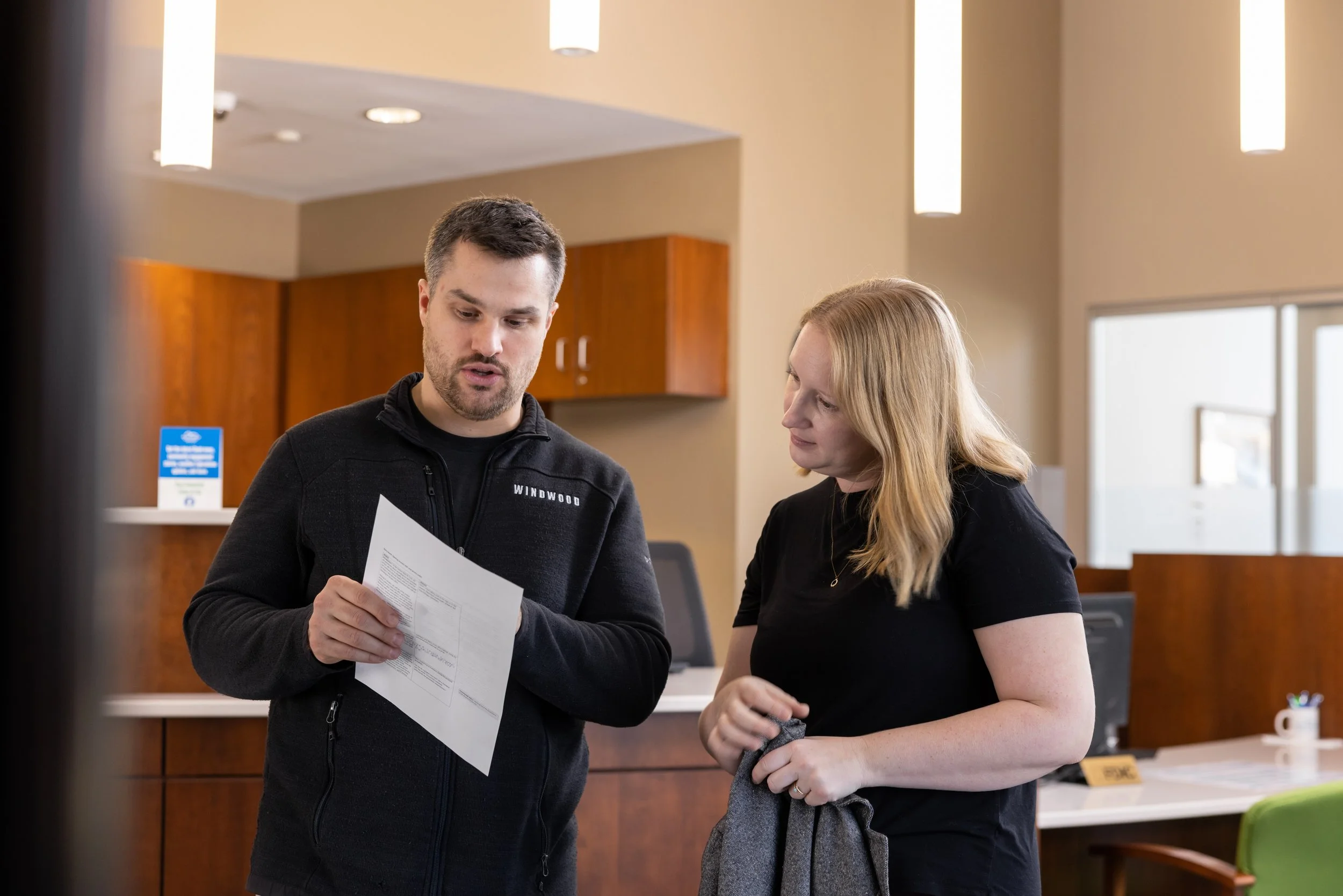 A man and woman standing at a reception desk indoors, looking at a piece of paper the man is holding.