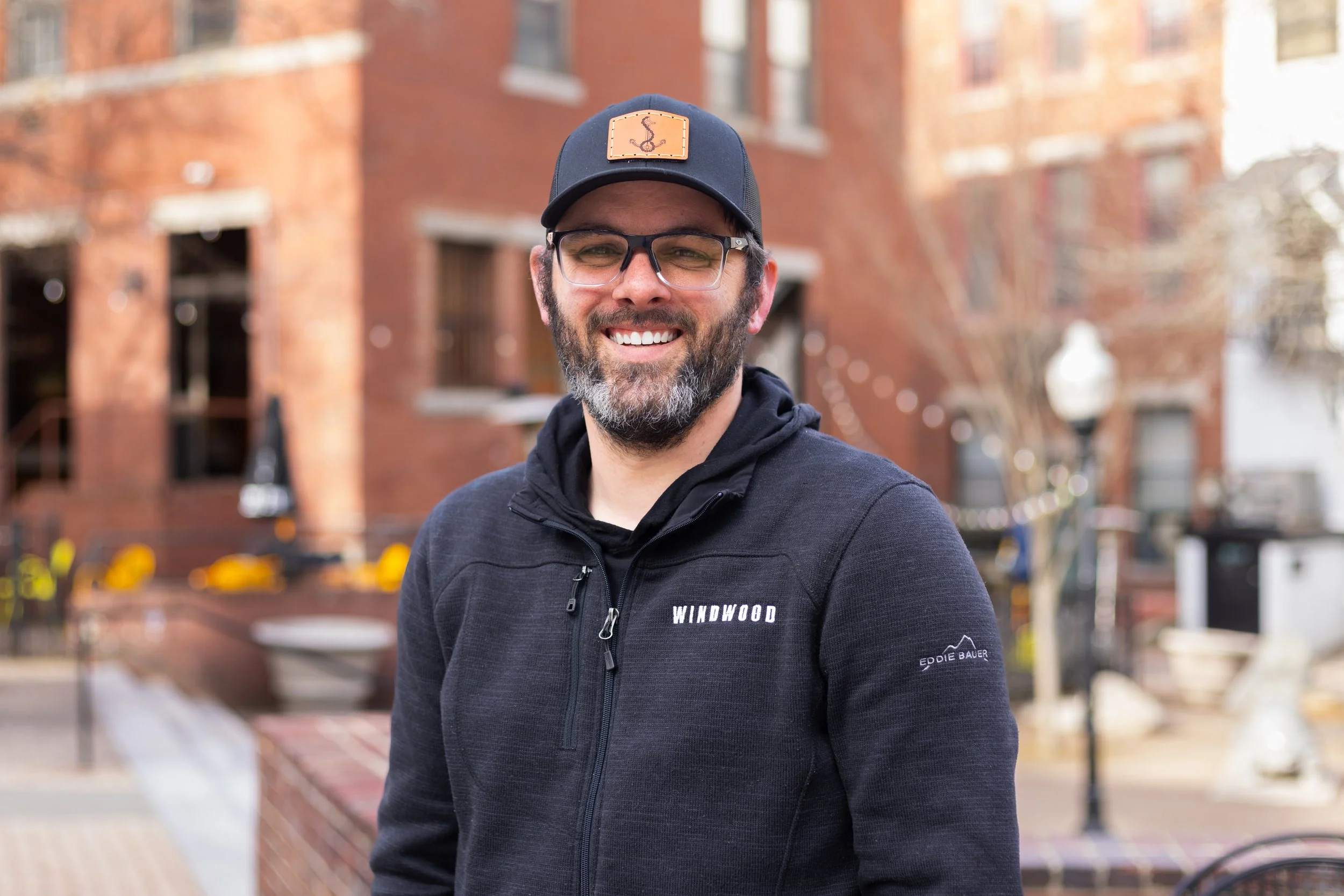 A smiling man with glasses, a beard, and a black cap stands outdoors in front of a brick building with trees and yellow flowers.