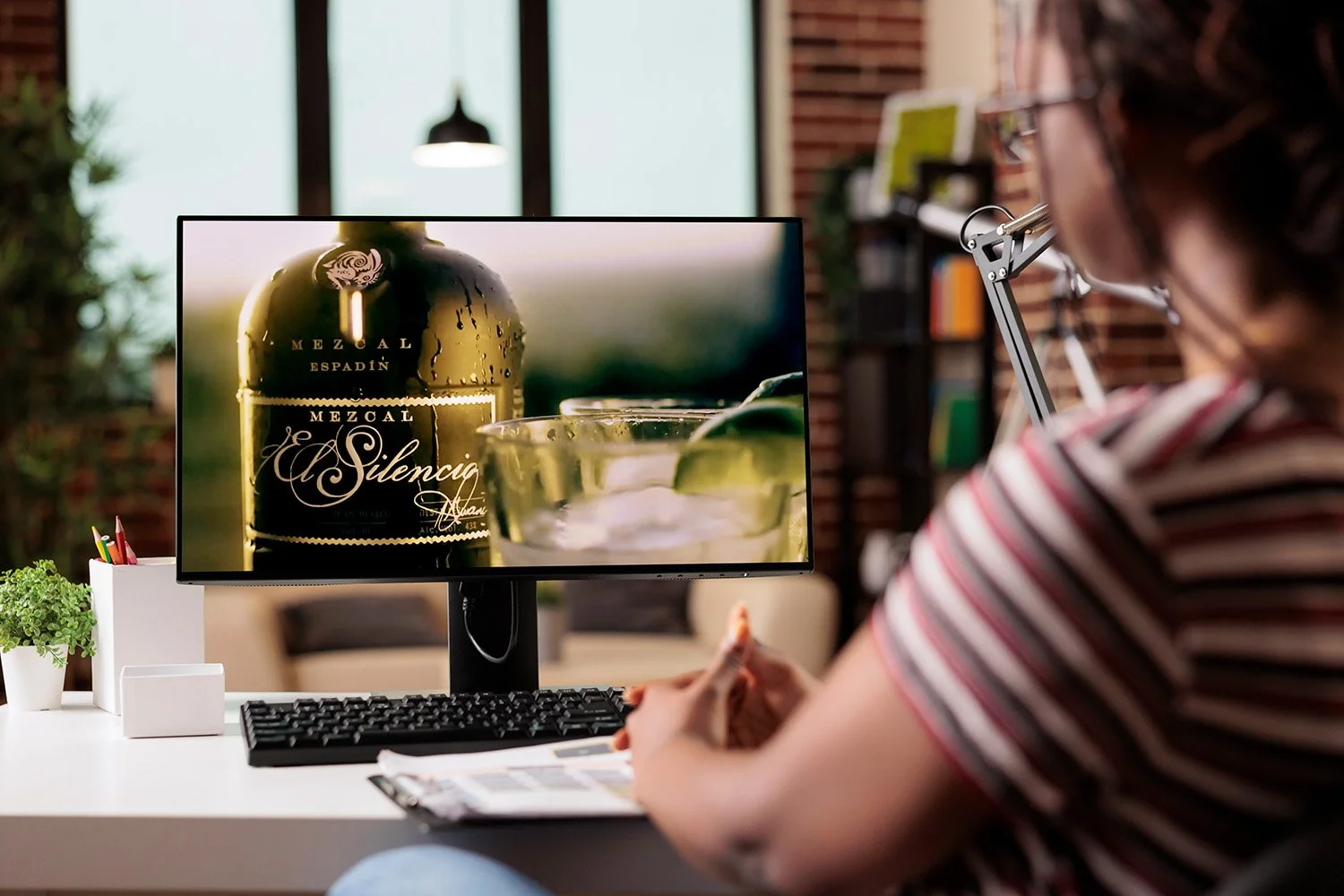A woman sitting at a desk, looking at a computer monitor displaying a bottle of mezcal and a glass with lime wedges, with a blurred background of a modern office or home space.
