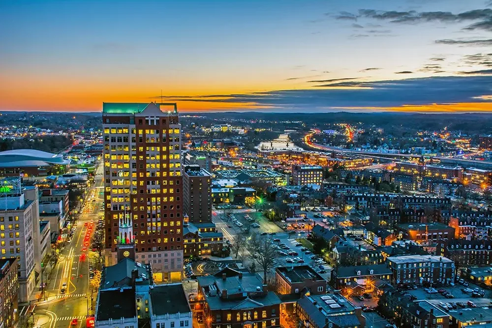 A Mancherster NH cityscape at dusk with a tall building illuminated from within and a sunset sky above, showing a mix of modern skyscrapers and smaller buildings, with a busy street grid below.