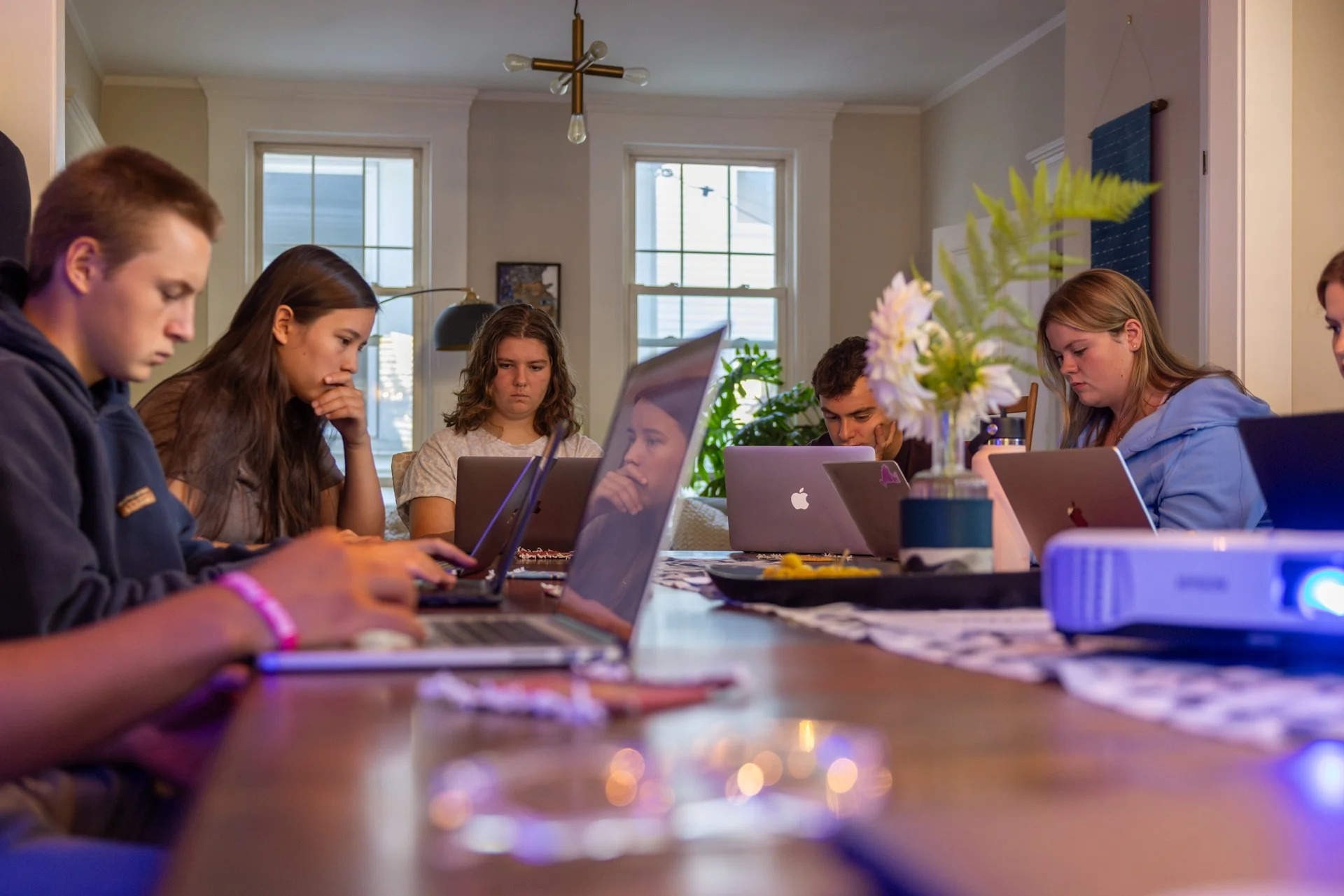 Windwood Productions video production meeting with a client at a shoot. Group of teenagers sitting at a dining table working on laptops, with a projector and flower vase in a well-lit dining room.