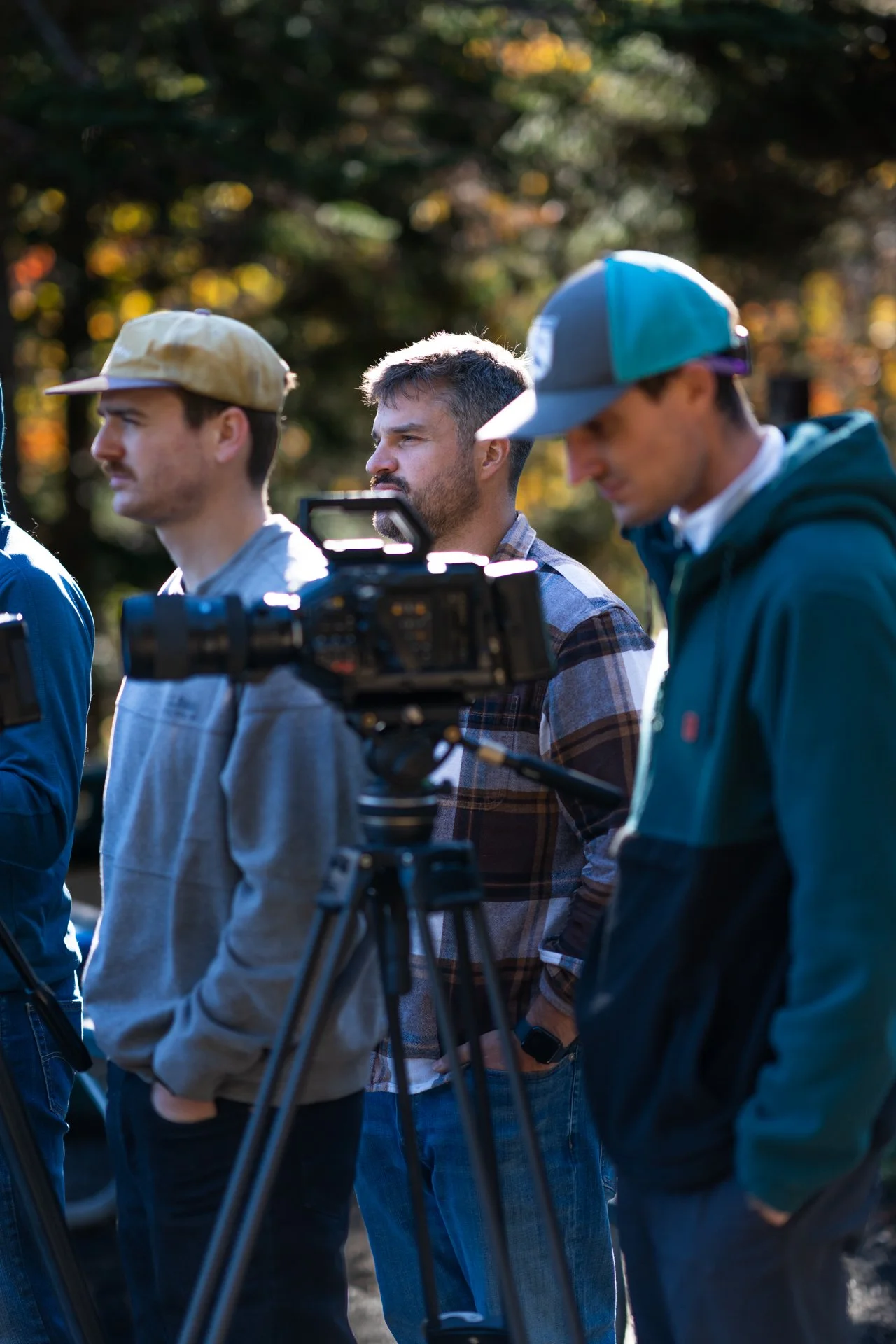 Group of three young men standing outdoors, two wearing baseball caps, one with a plaid shirt, and one in a hoodie, near a camera mounted on a tripod, with trees and sunlight in the background.