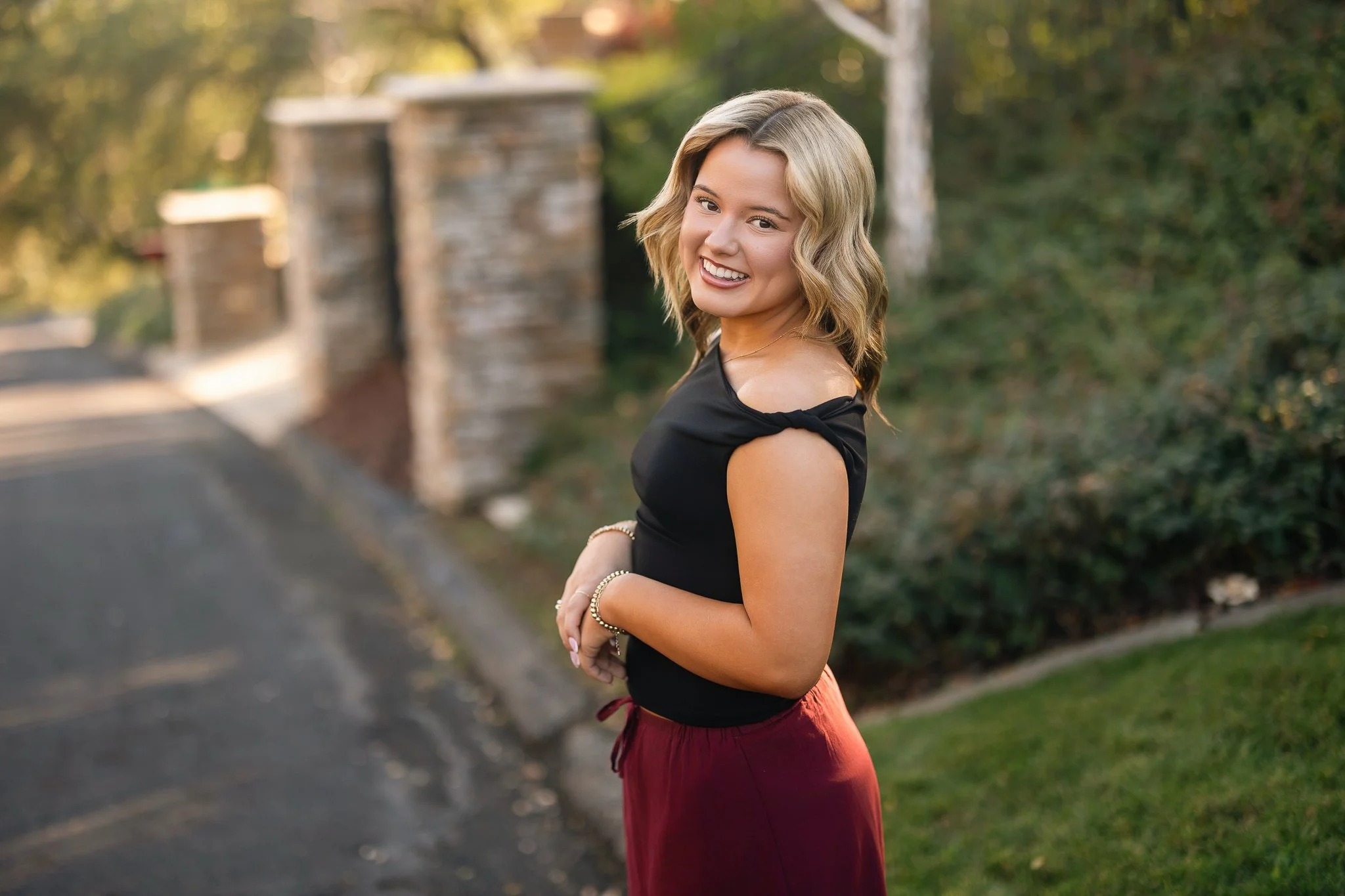 A young woman smiling outdoors in a black top and red pants, standing near a sidewalk with greenery and blurred trees in the background during sunset.