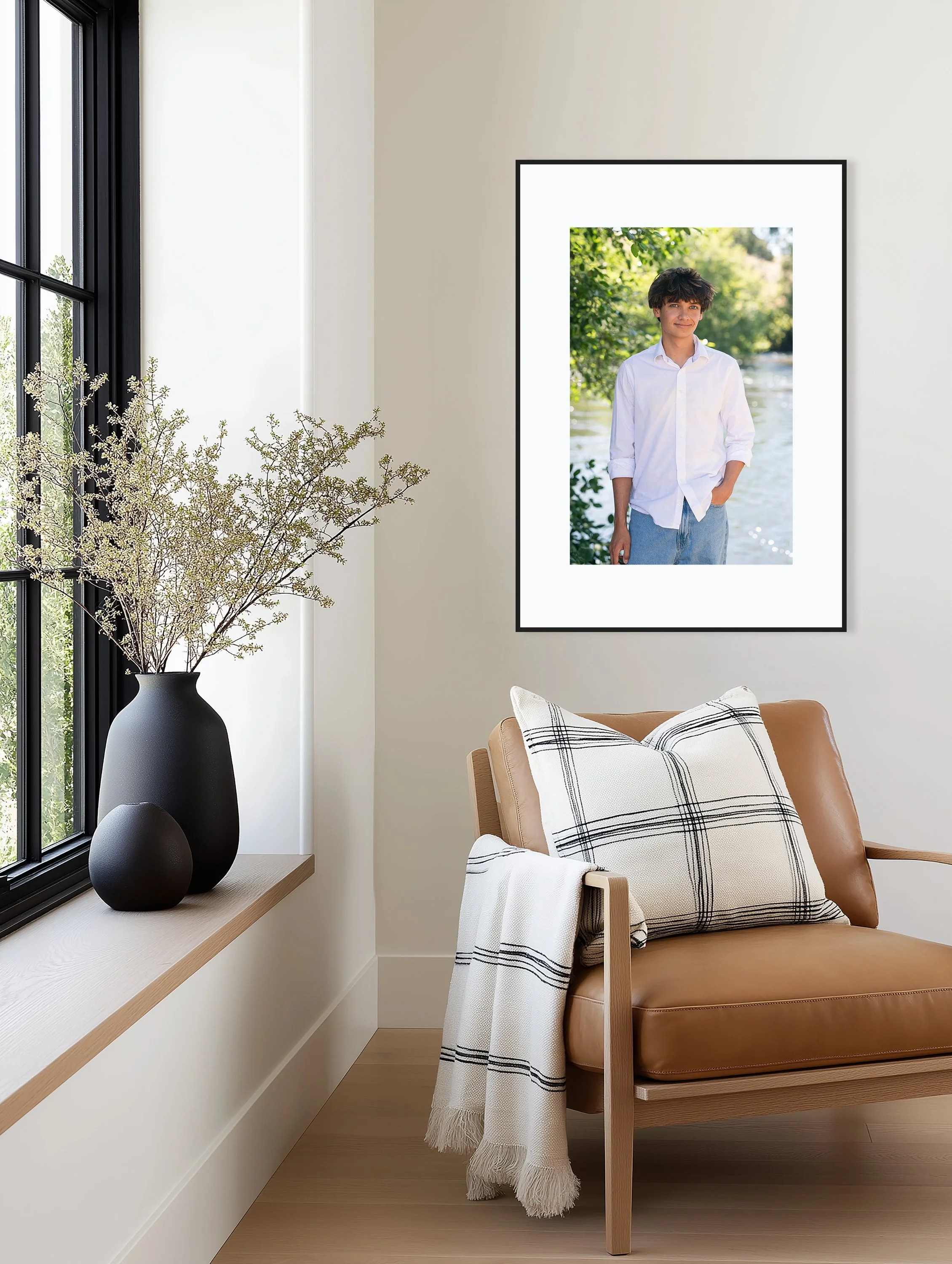 Interior of a modern room with a framed photo of a young man in a white shirt and blue jeans on the wall, a window with black frames, a wooden armchair with a beige cushion and a white throw blanket with black stripes, and a black vase with dried flowers on the window sill.