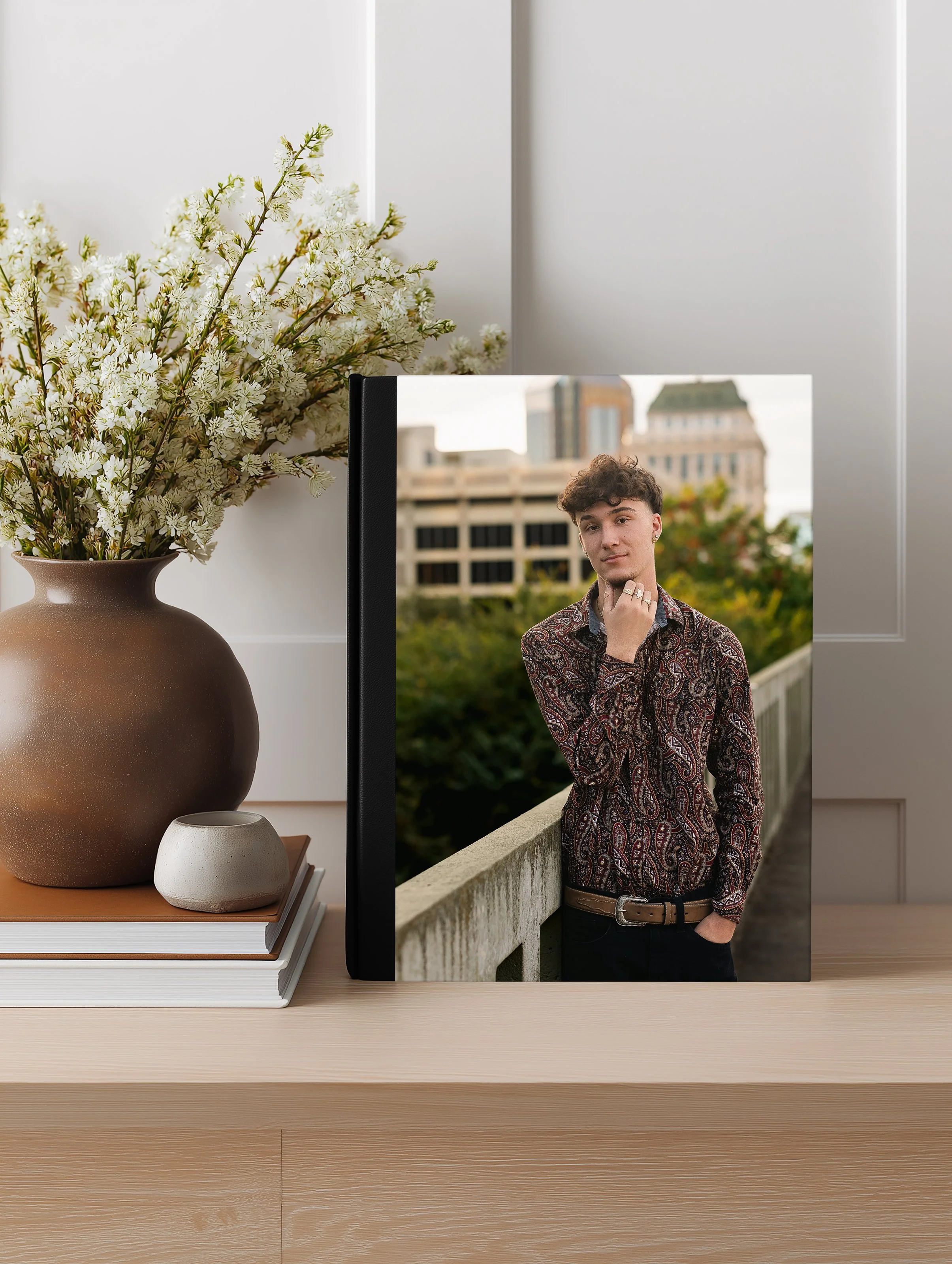 A large brown vase filled with white flowering branches on a beige wooden table, partially obscuring a photograph of a young man with curly hair and a patterned shirt standing outdoors near a concrete railing, with buildings and trees in the background.