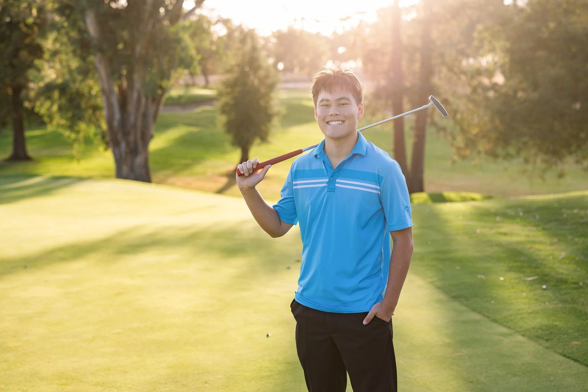 Senior guy in a blue polo holding a putter over his shoulder on a golf course at sunset, golf senior pictures in Sacramento