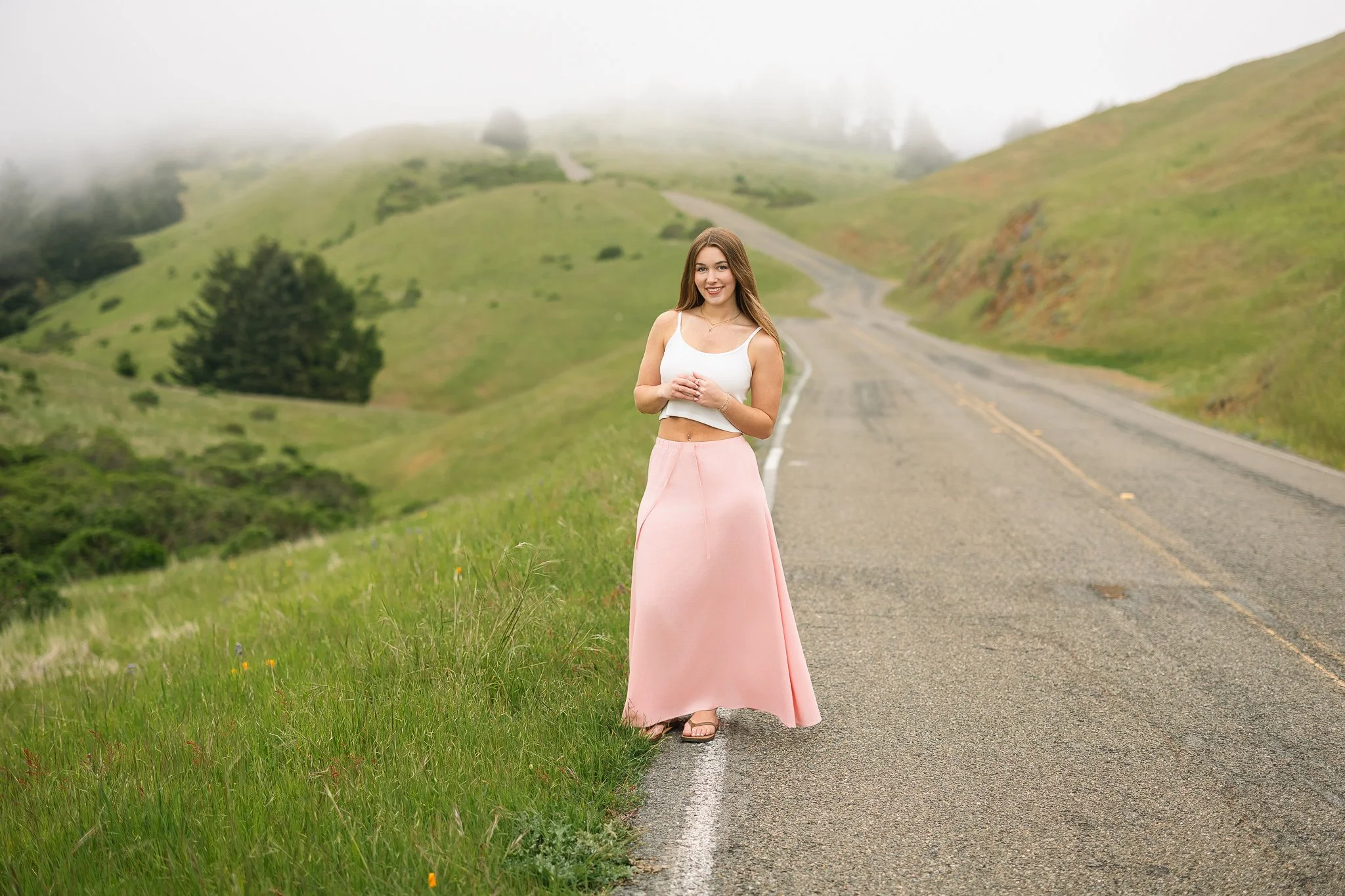Granite Bay High School senior in a white tank top and long pink skirt standing beside a winding road at Mount Tamalpais, surrounded by green hills and rolling fog.