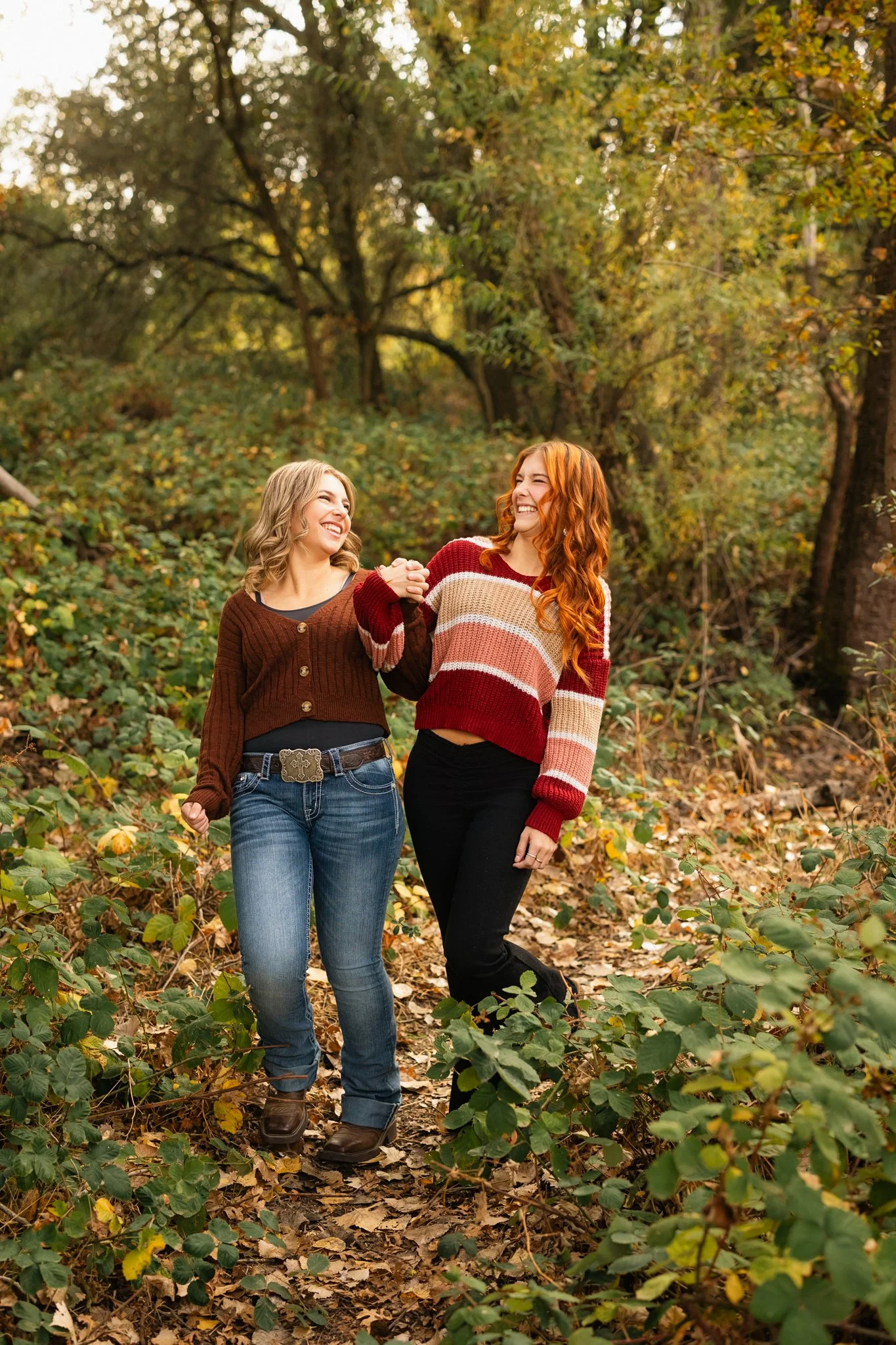 Two women walking in a wooded area during autumn, holding hands and smiling at each other.