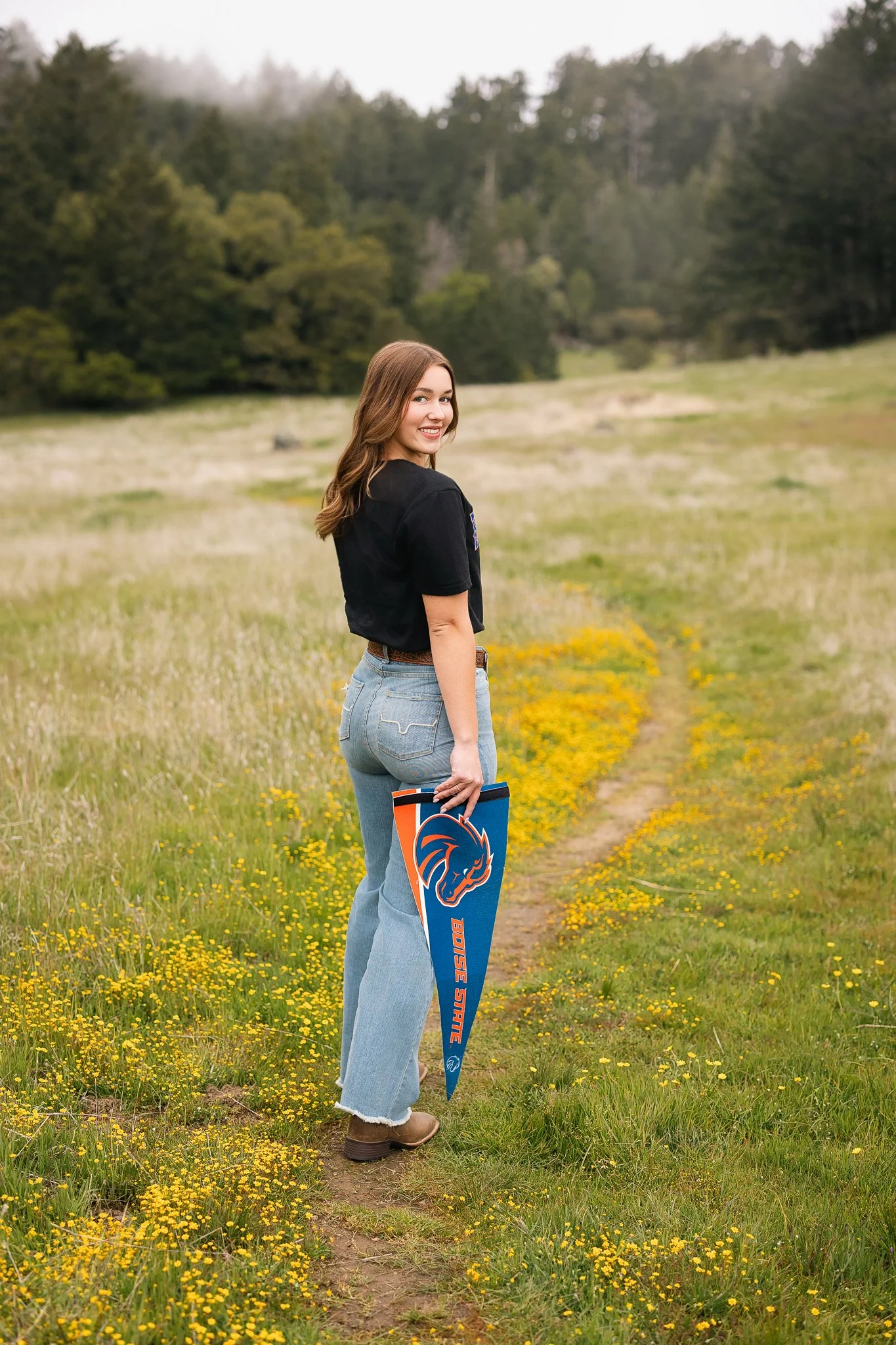  Granite Bay High School senior holding a Boise State pennant and smiling over her shoulder on a Mount Tamalpais trail lined with yellow wildflowers. 