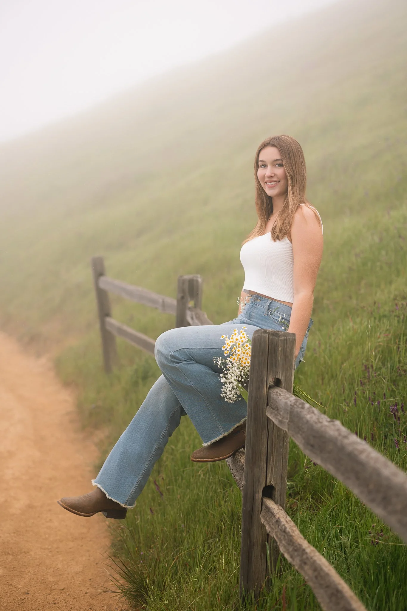  Graduating senior sitting on a wooden fence at Mount Tamalpais, wearing a white tank top, jeans, and brown boots, with a bouquet of daisies beside her in the fog. 