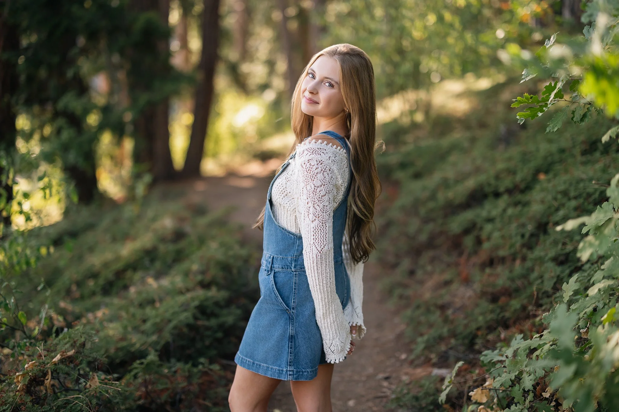 Young woman standing on a nature trail in a wooded area, smiling, wearing a white crocheted top under denim overalls, with long brown hair, surrounded by green leaves and sunlight filtering through trees.