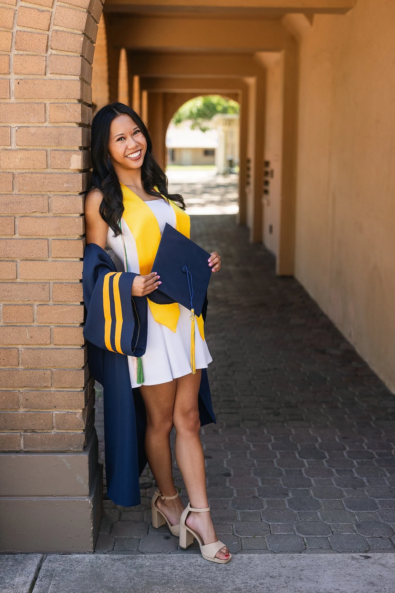 A young woman in a graduation gown and cap, smiling, leaning against a brick wall in a corridor, holding her cap in her right hand, with a background of trees and a building.