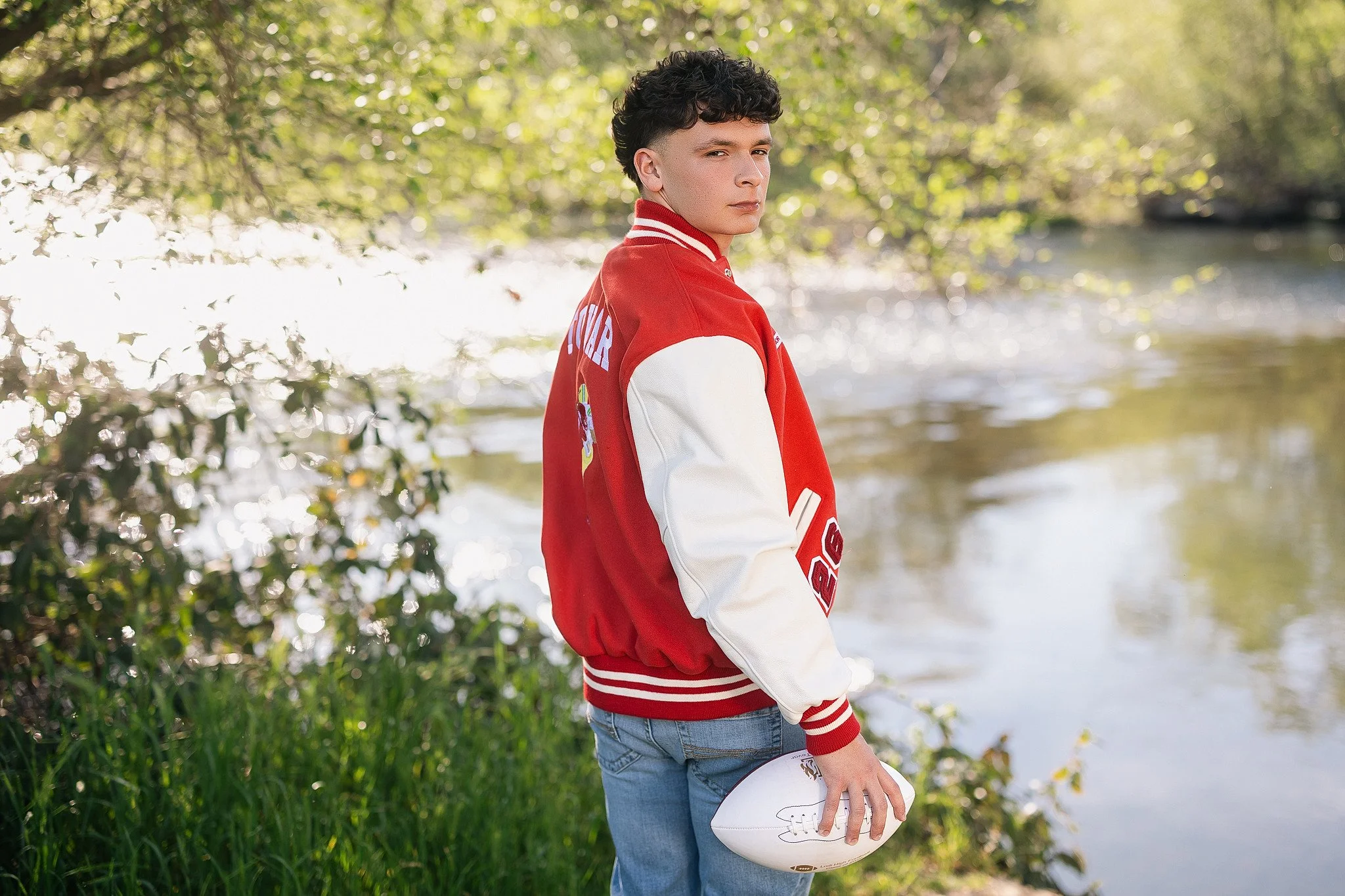 Lodi High School senior guy wearing his letterman jacket and holding a football during his outdoor senior pictures along the Mokelumne River in Lodi, California.