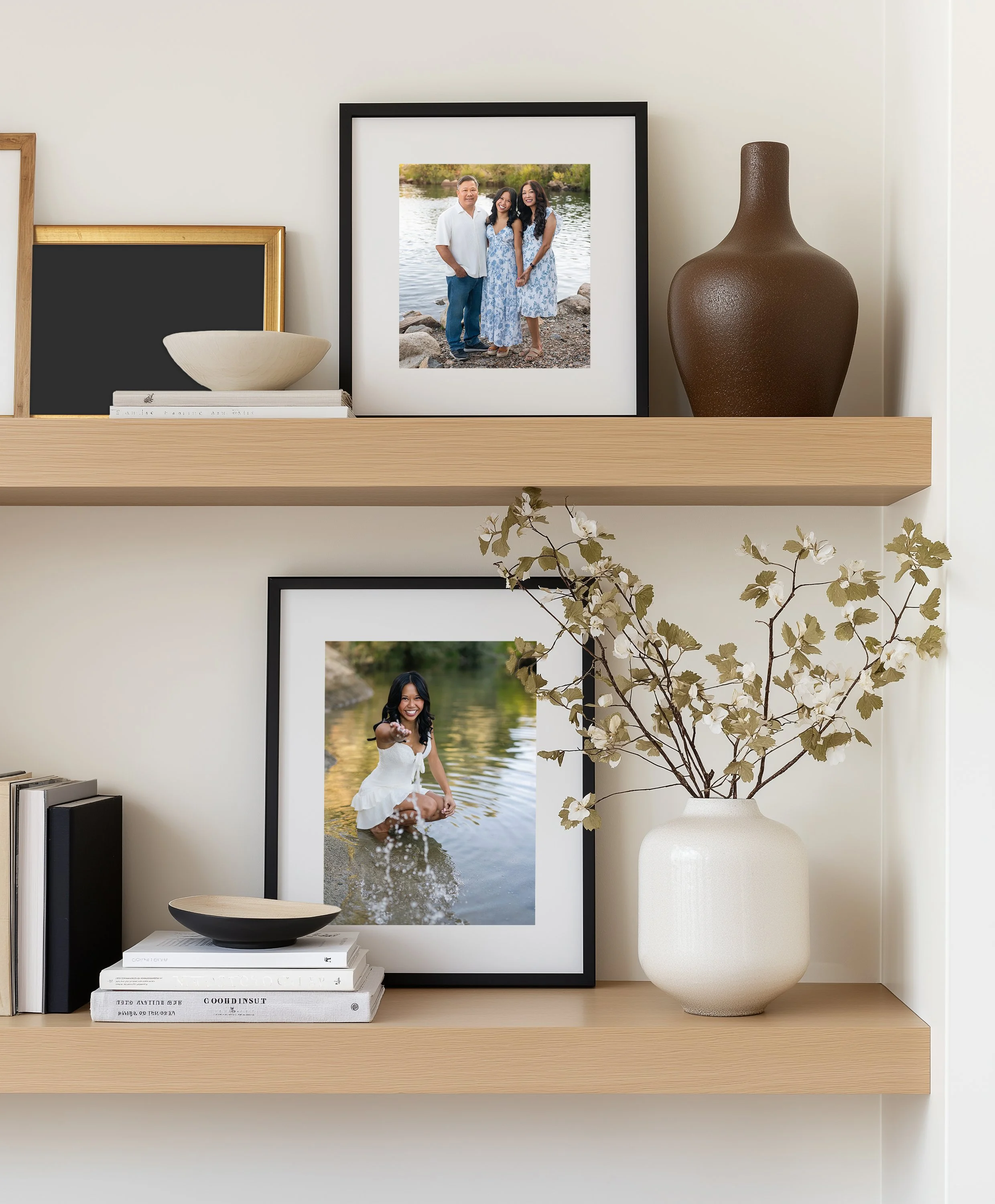 A wooden shelf displaying a framed photograph of three people by a body of water, with a large brown textured vase, a smaller black and white book, a large black bowl, and some stacked white books underneath.