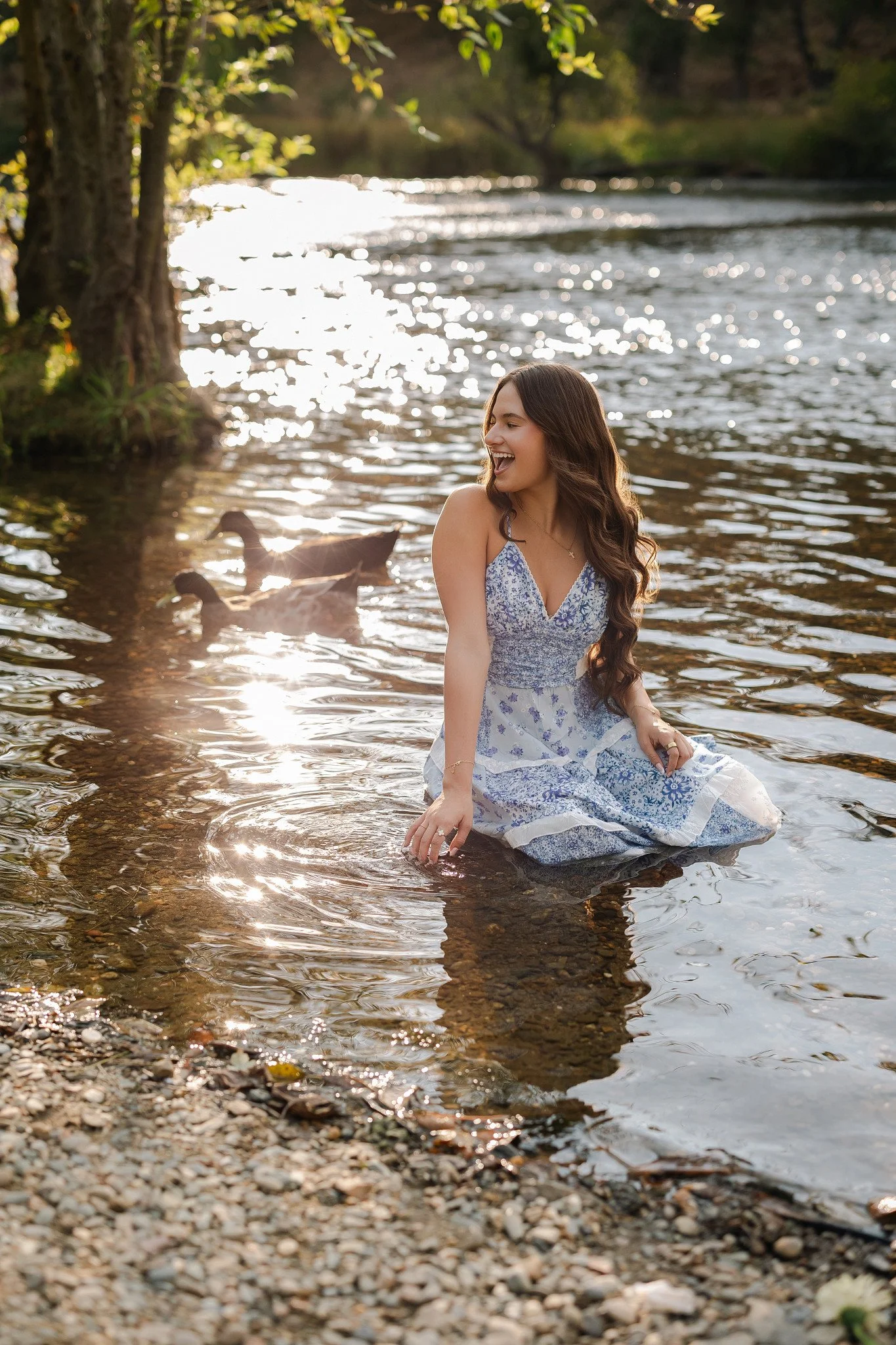 Lodi High School senior laughing while sitting in the shallow Mokelumne River in a blue floral dress, with sunlight sparkling on the water and ducks swimming behind her.