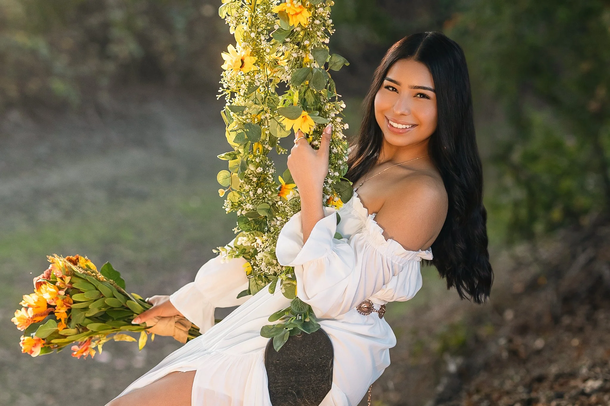 A woman with long black hair and a white off-the-shoulder dress smiling and holding a floral wreath of yellow and orange flowers on a swing decorated with flowers, in a natural outdoor setting.