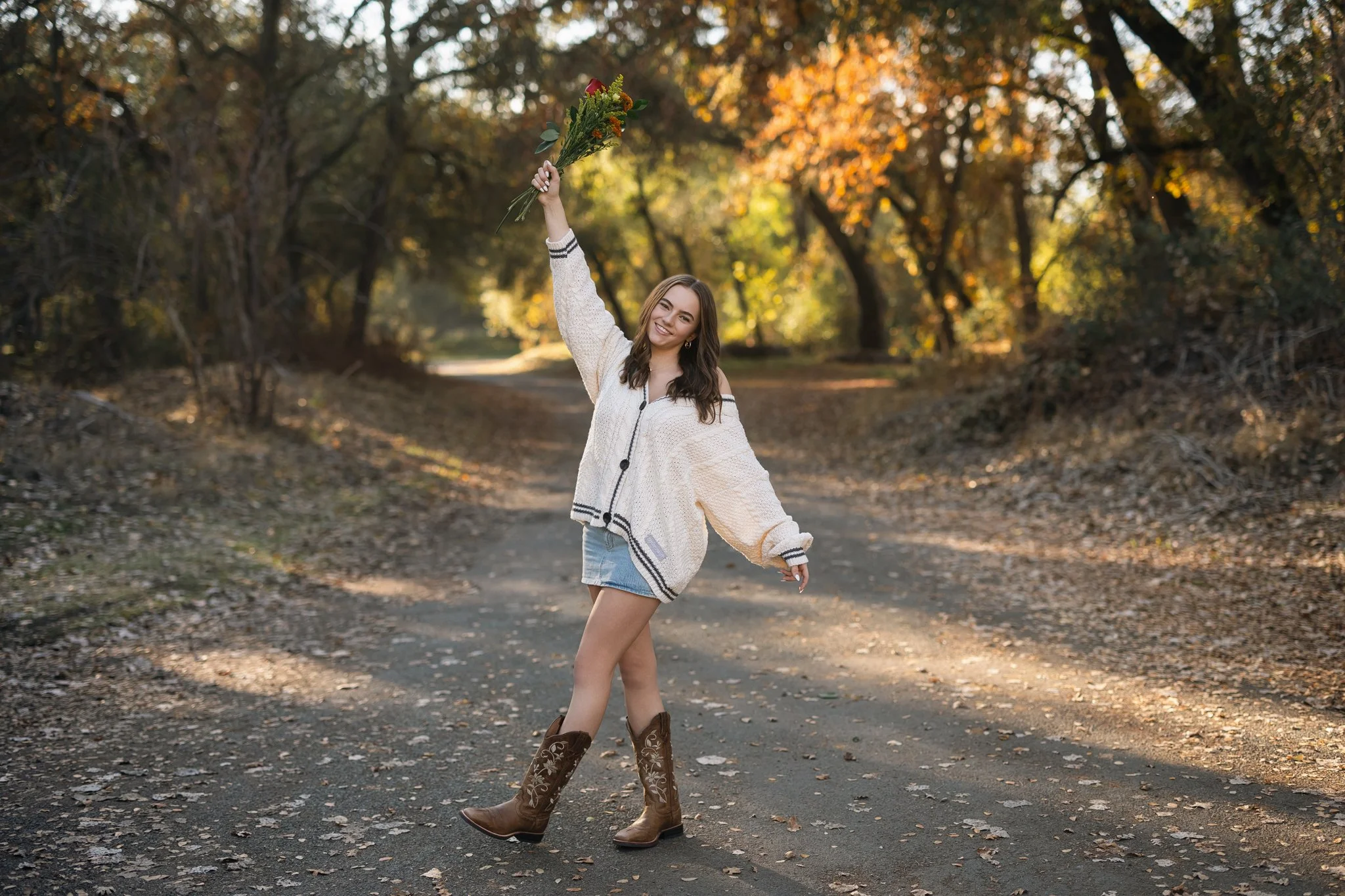 Fall senior pictures of a girl wearing denim skirt, taylor swift sweater and cowboy boots holding flowers on a road near Folsom Lake