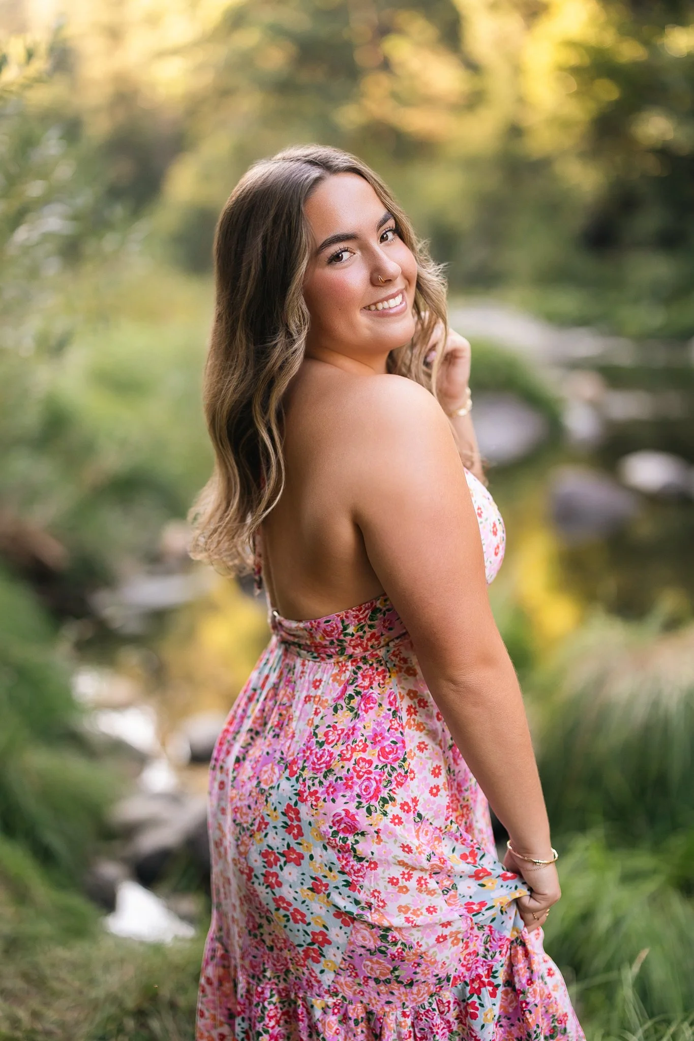 A smiling young woman with long, wavy brown hair, standing outdoors in a natural setting with blurred greenery and a small stream in the background. She is wearing a sleeveless, floral pink and white dress and has a nose ring and bracelet.