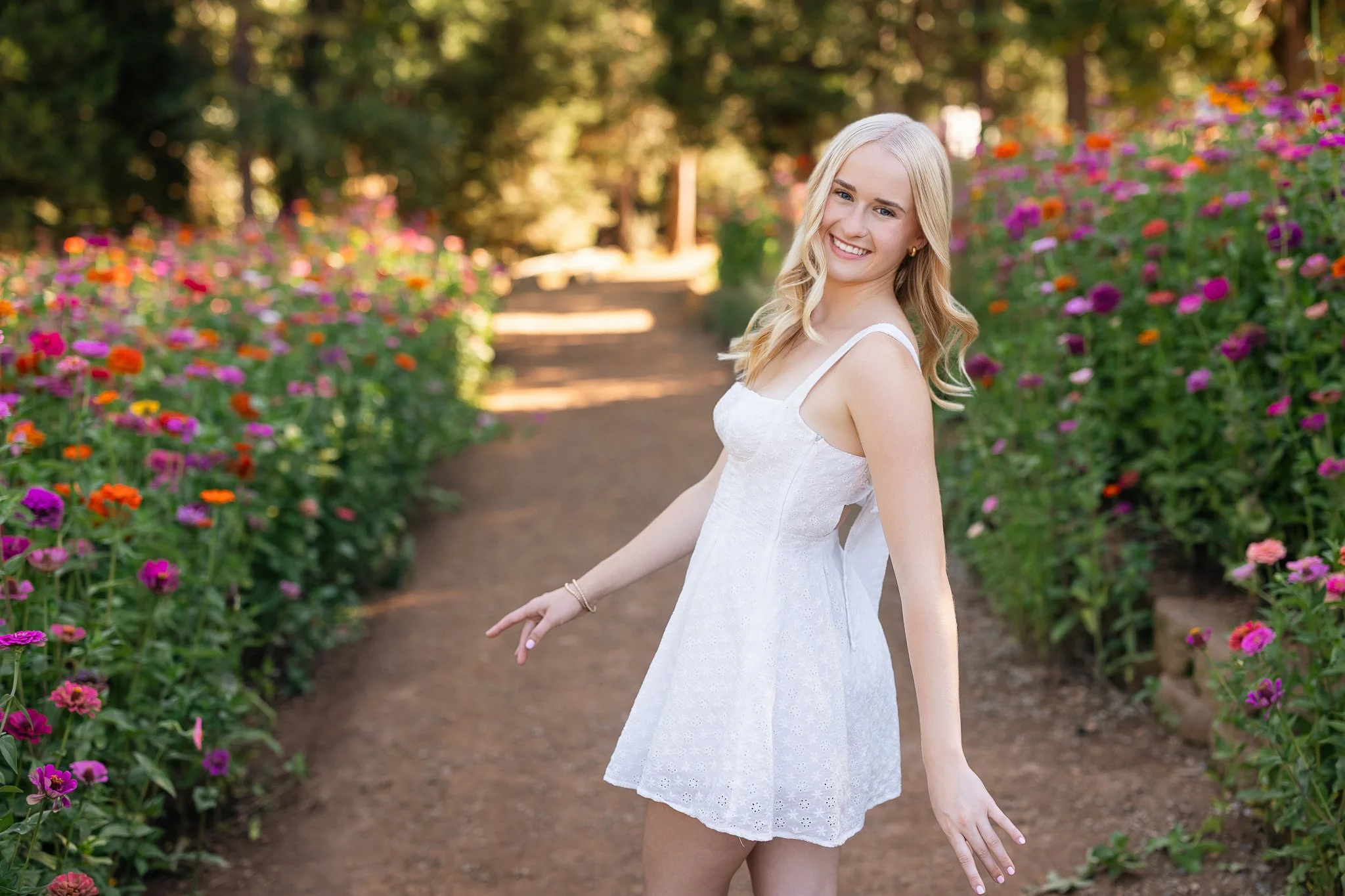 A young woman in a white dress smiling and posing on a flower-lined pathway in a park with trees in the background.
