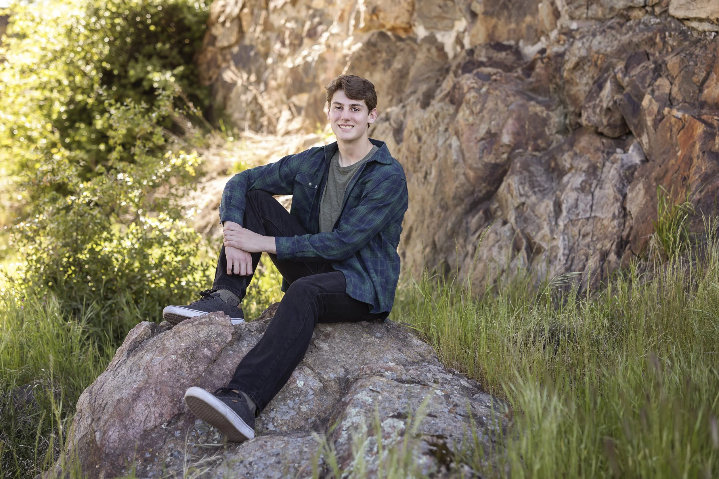 A young man with short brown hair smiling, sitting on a rock in front of a rocky hillside, wearing a dark plaid shirt and dark pants, with greenery and trees in the background on a sunny day.