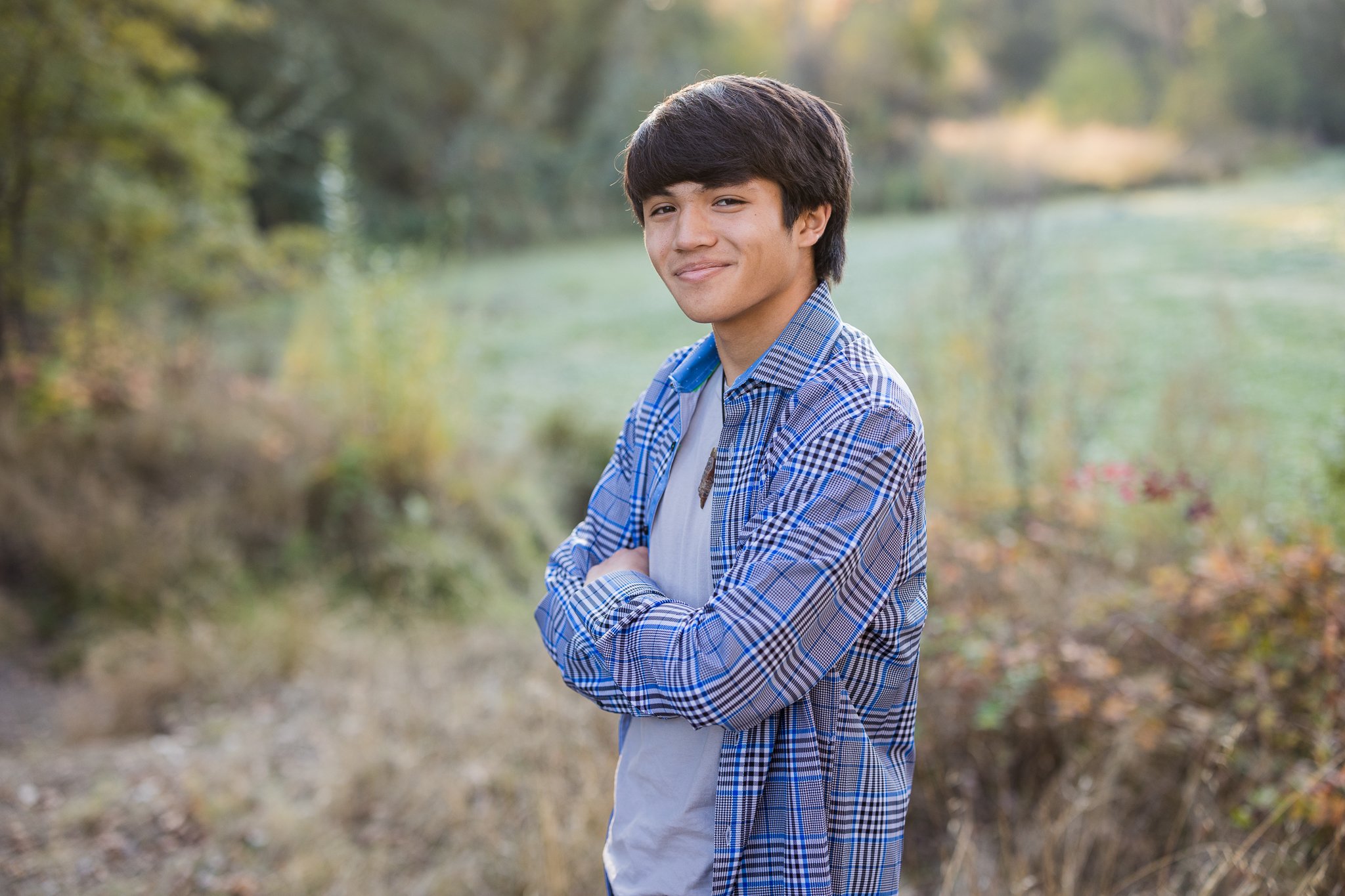 A young man with dark hair, wearing a blue checkered shirt and gray t-shirt, standing outdoors in a park or natural setting with blurred trees and greenery in the background.
