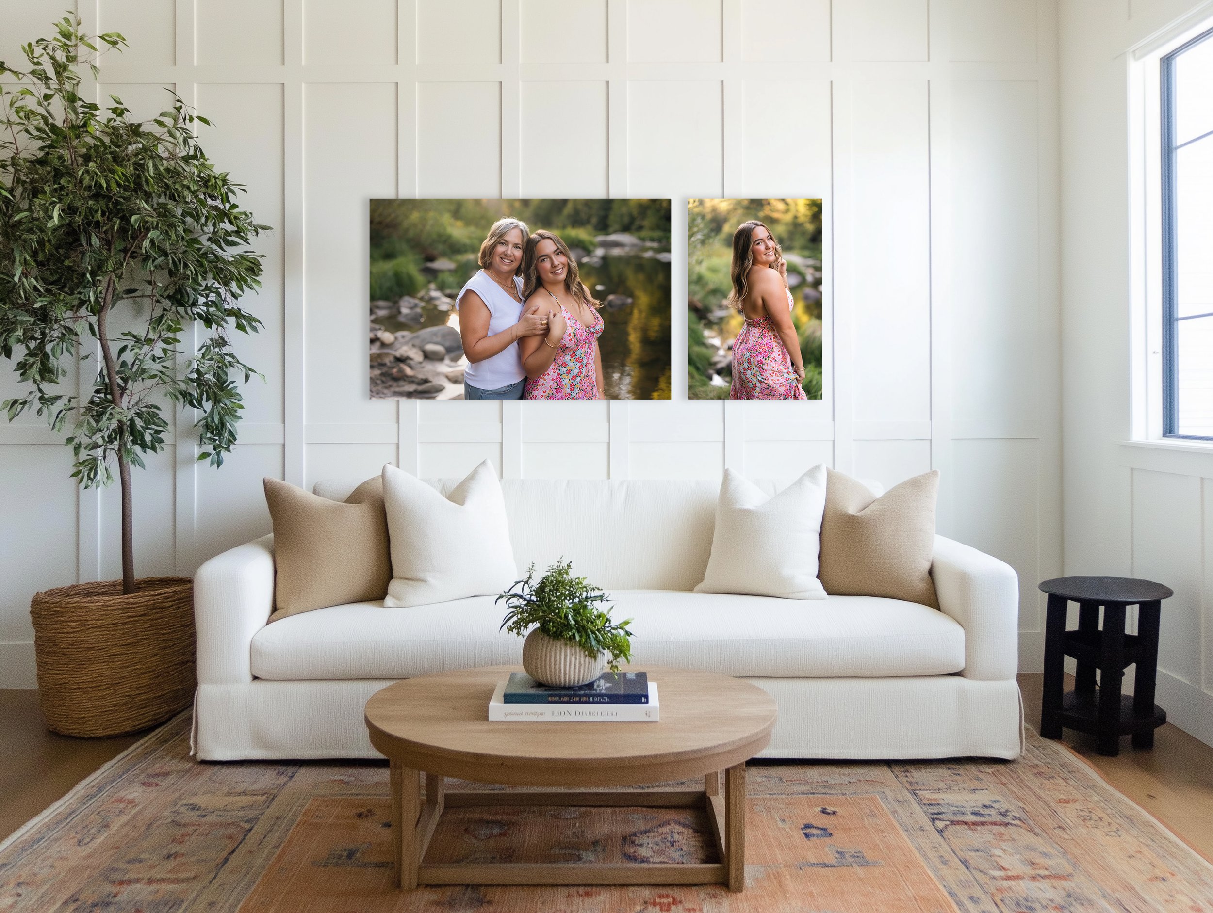 Living room with white sofa, beige pillows, wooden coffee table with a plant and books, a large potted plant on the left, and two framed photos of women outdoors on white paneled wall, next to a window.