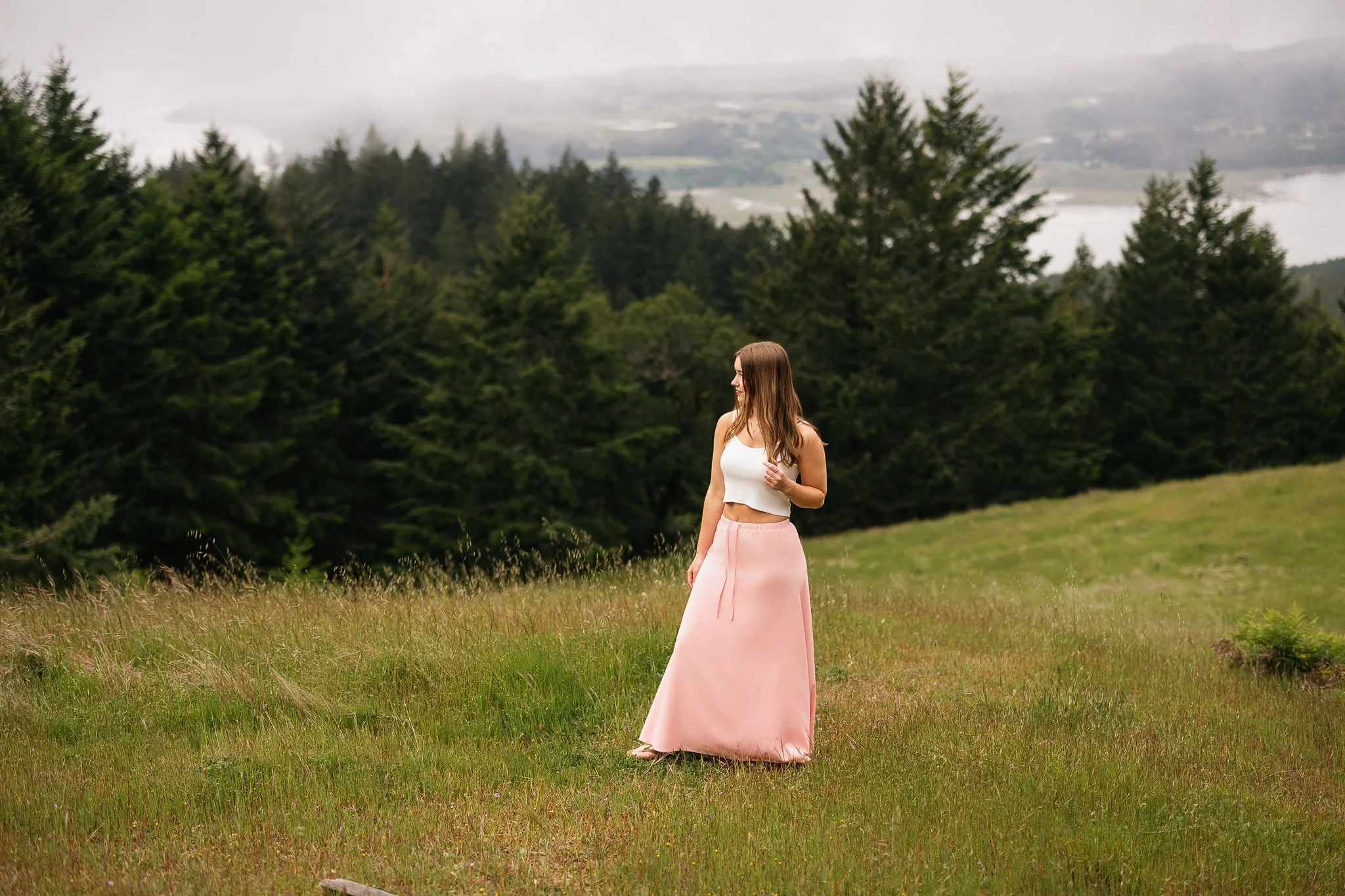 High school senior girl wearing a pink flowy skirt and white top standing on  a mountain over looking stinson beach