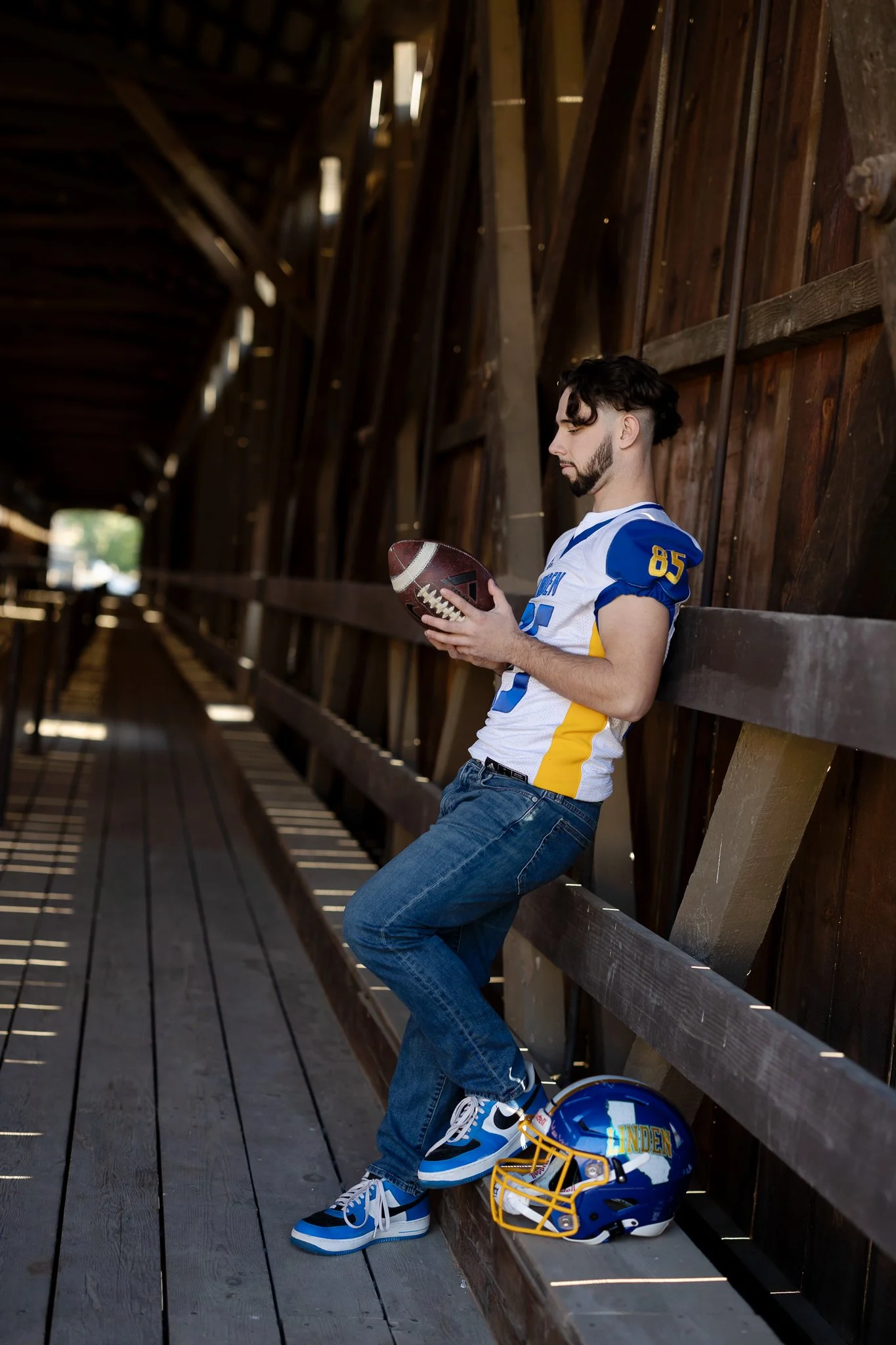 Linden High School senior football player holding a football inside the historic covered bridge at Knights Ferry, with his Linden football helmet beside him.