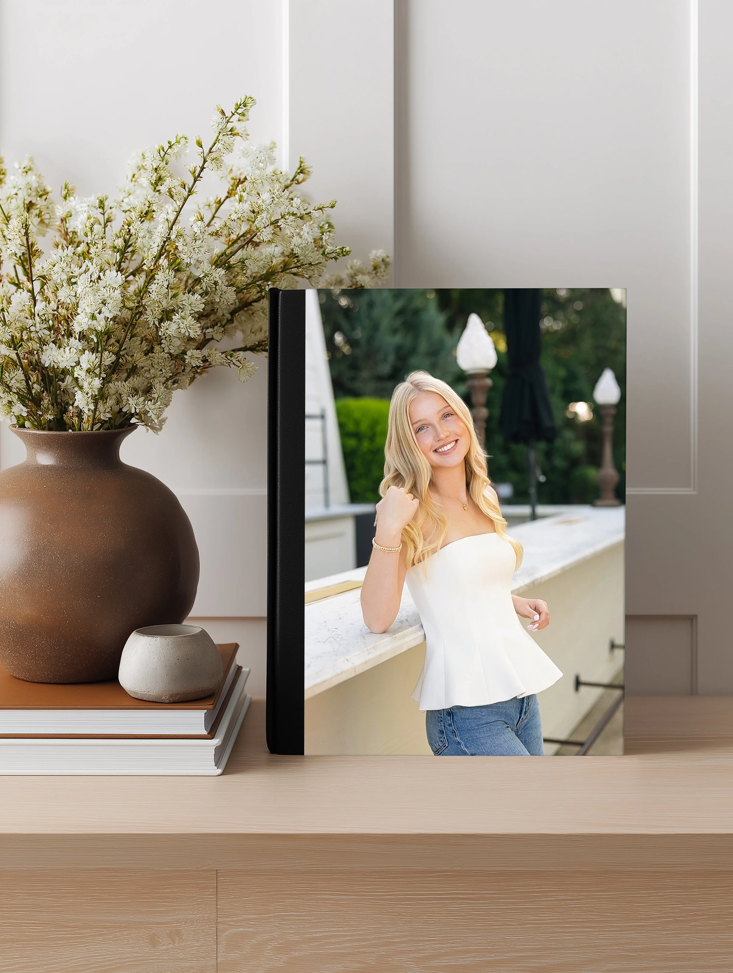 A desk with a stack of books, a large brown vase with white flowers, a small gray vase, and a framed photo of a young blonde woman smiling outdoors.