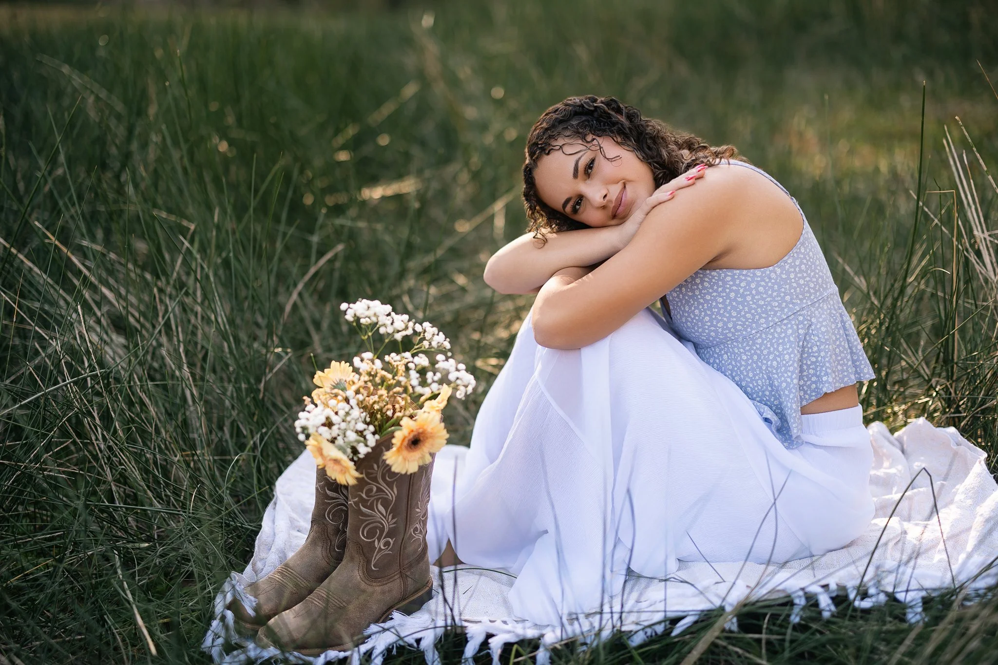 A woman with curly hair lying on her side with her arms around her knee, smiling softly. She is wearing a light purple dress with small white patterns and sitting outdoors among tall grass, near a small bouquet of yellow and white flowers placed on a white cloth.