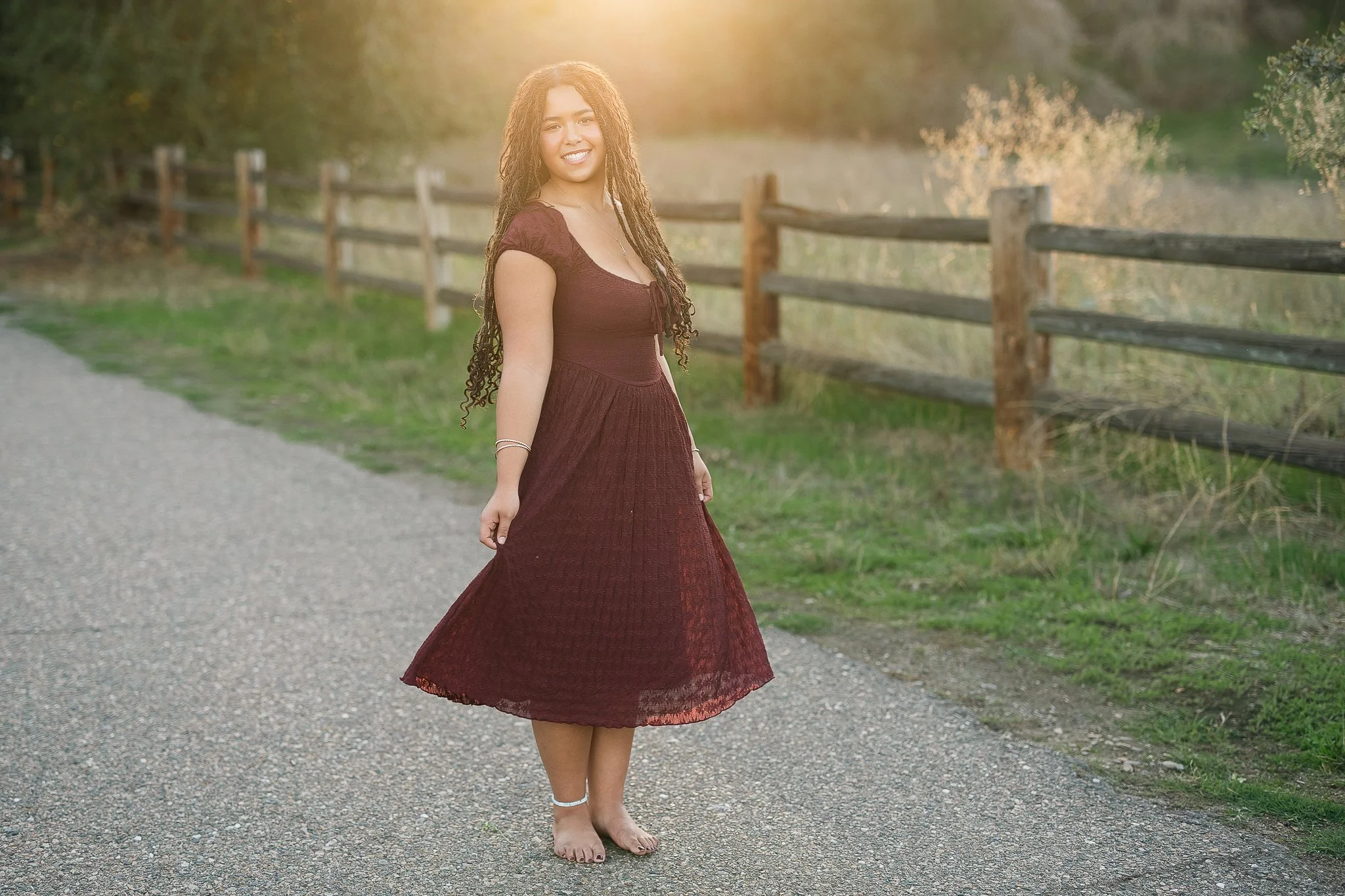 Oak Ridge High School senior girl in a burgundy dress walking barefoot along a rustic split-rail fence at golden hour, photographed by Folsom Lake senior photographer
