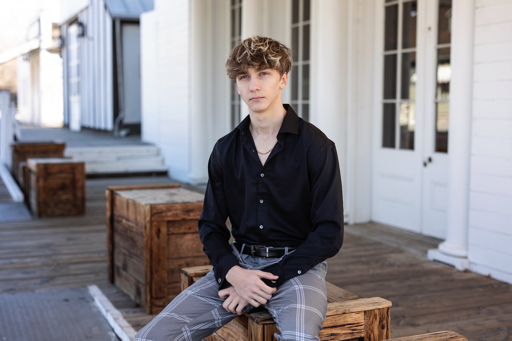 A young man with curly hair wearing a black shirt and plaid pants sitting on a wooden bench outside a house with white siding and large windows.