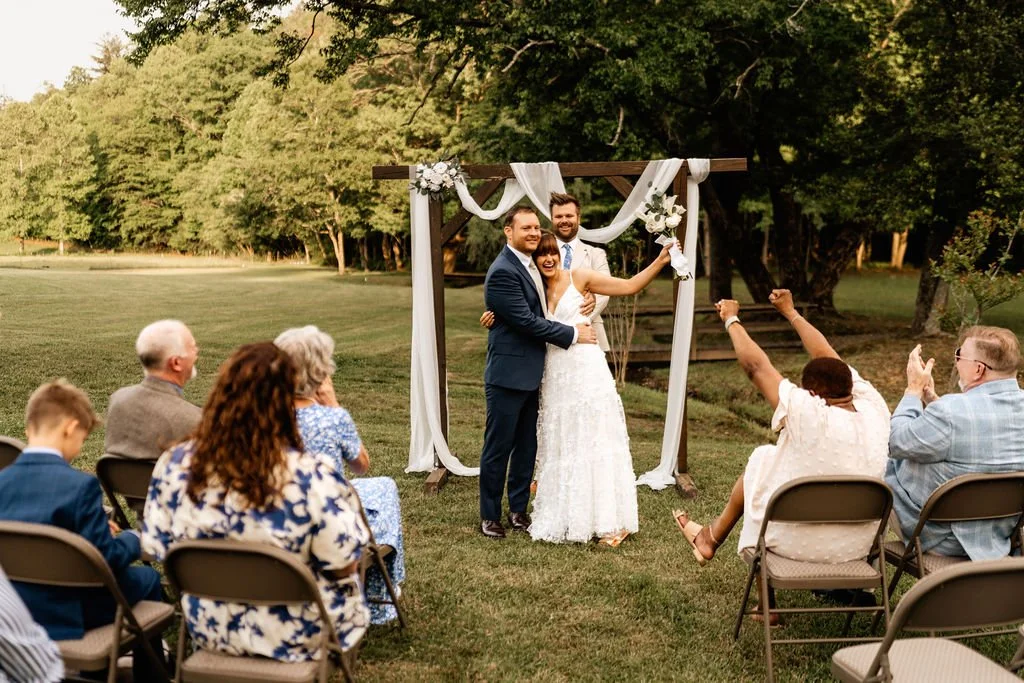 Newlyweds celebrating with cheering guests after their outdoor ceremony on a   green meadow at a Blue Ridge, Georgia barn wedding venue