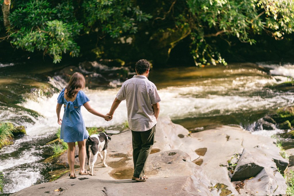 A couple and their dog walking together along a riverbank, enjoying the breathtaking mountain scenery