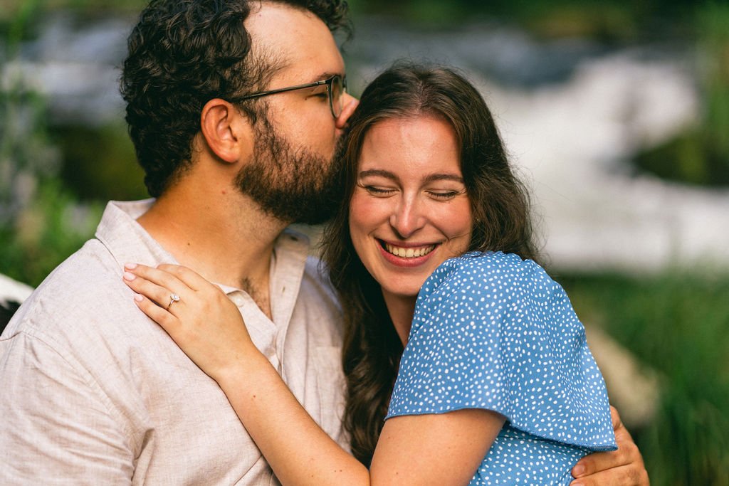 One partner kissing the other’s forehead, with the engagement ring beautifully displayed.