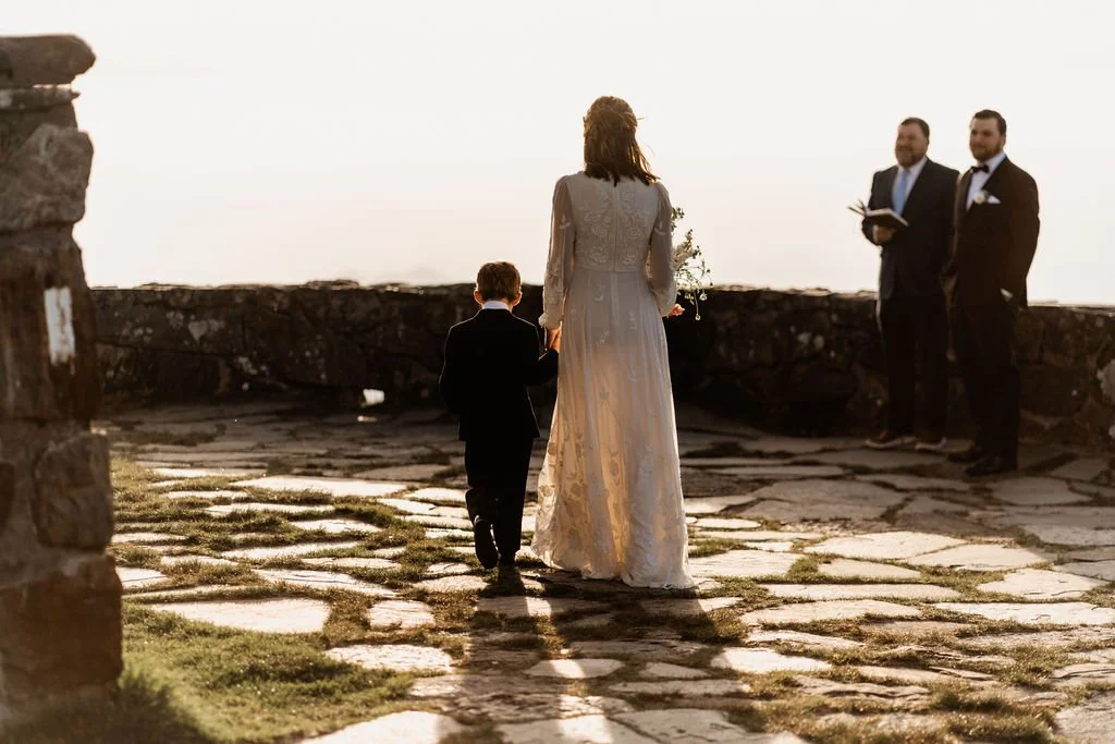 Bride walking across the stone terrace at Wayah Bald toward their officiant with the groom waiting during their intimate mountain elopement ceremony, Nantahala National Forest, North Carolina