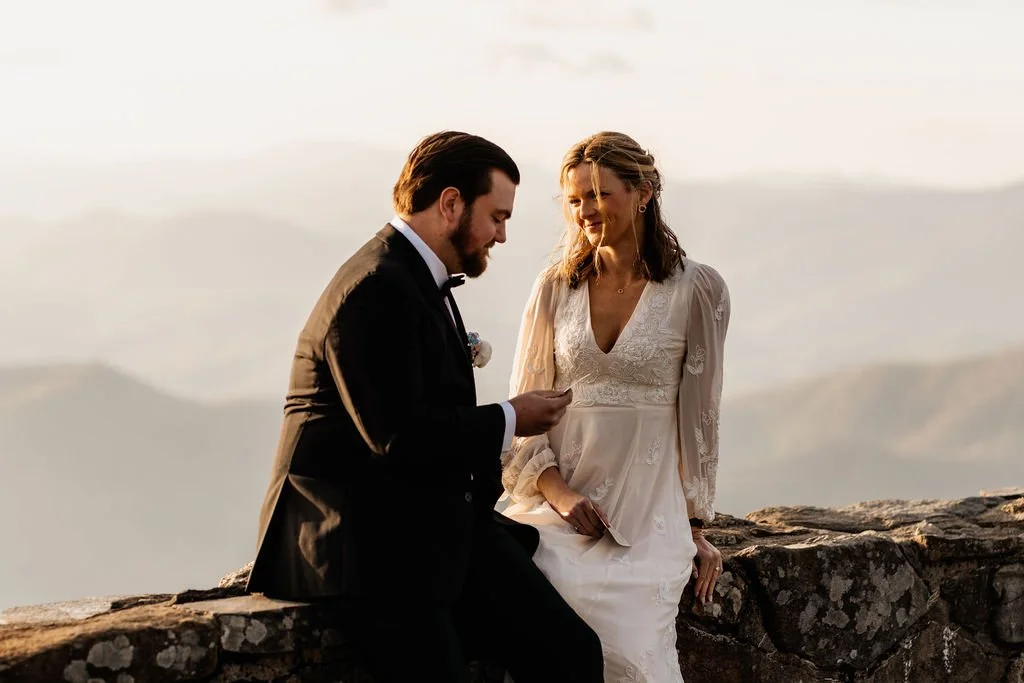 Bride and groom seated together on the stone wall at Wayah Bald sharing an intimate moment during their ceremony, misty Appalachian mountain ridges in the background
