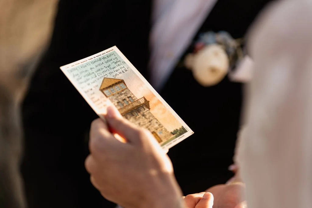 Close-up of bride's hands holding a vintage Wayah Bald postcard with handwritten love note during intimate mountain elopement ceremony, Nantahala National Forest NC