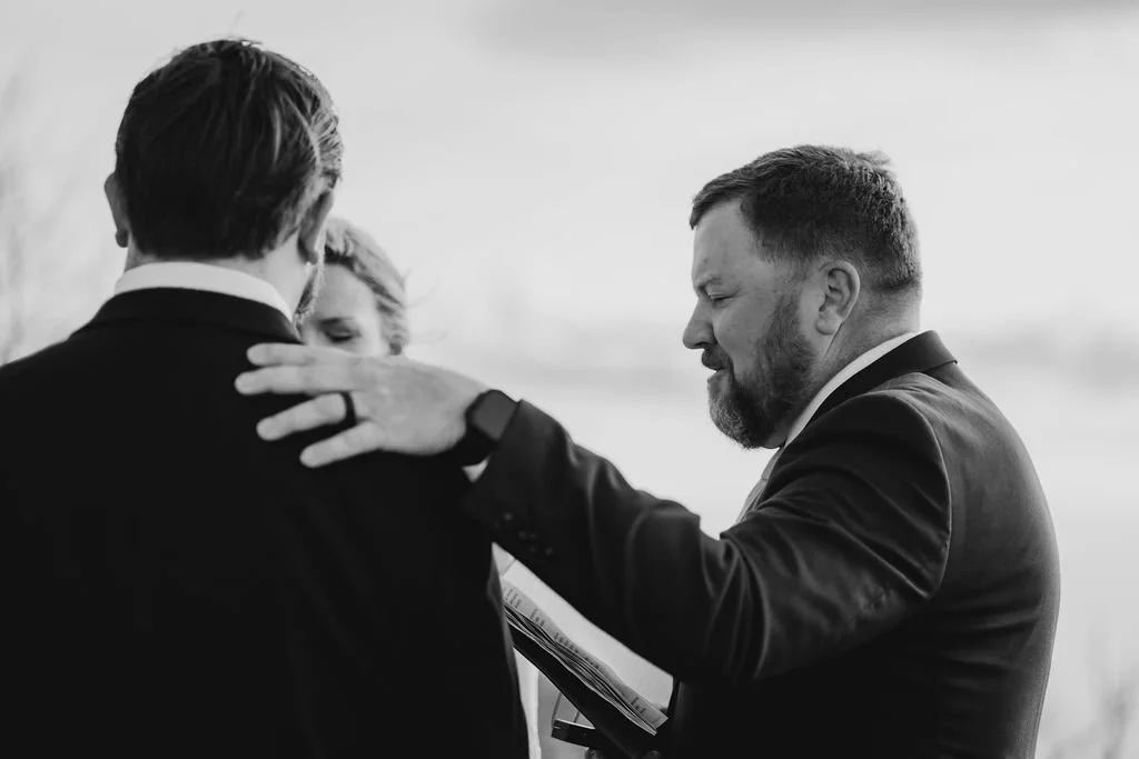 Black and white photo of officiant placing hand on groom's shoulder in a tender blessing moment during intimate Appalachian mountain wedding ceremony at Wayah Bald